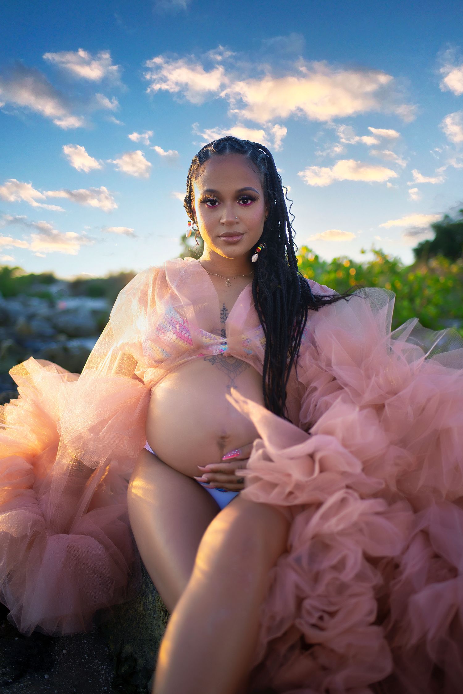 Pregnant woman in elegant pink ruffled tulle maternity gown cradling baby bump outdoors under a blue sky.