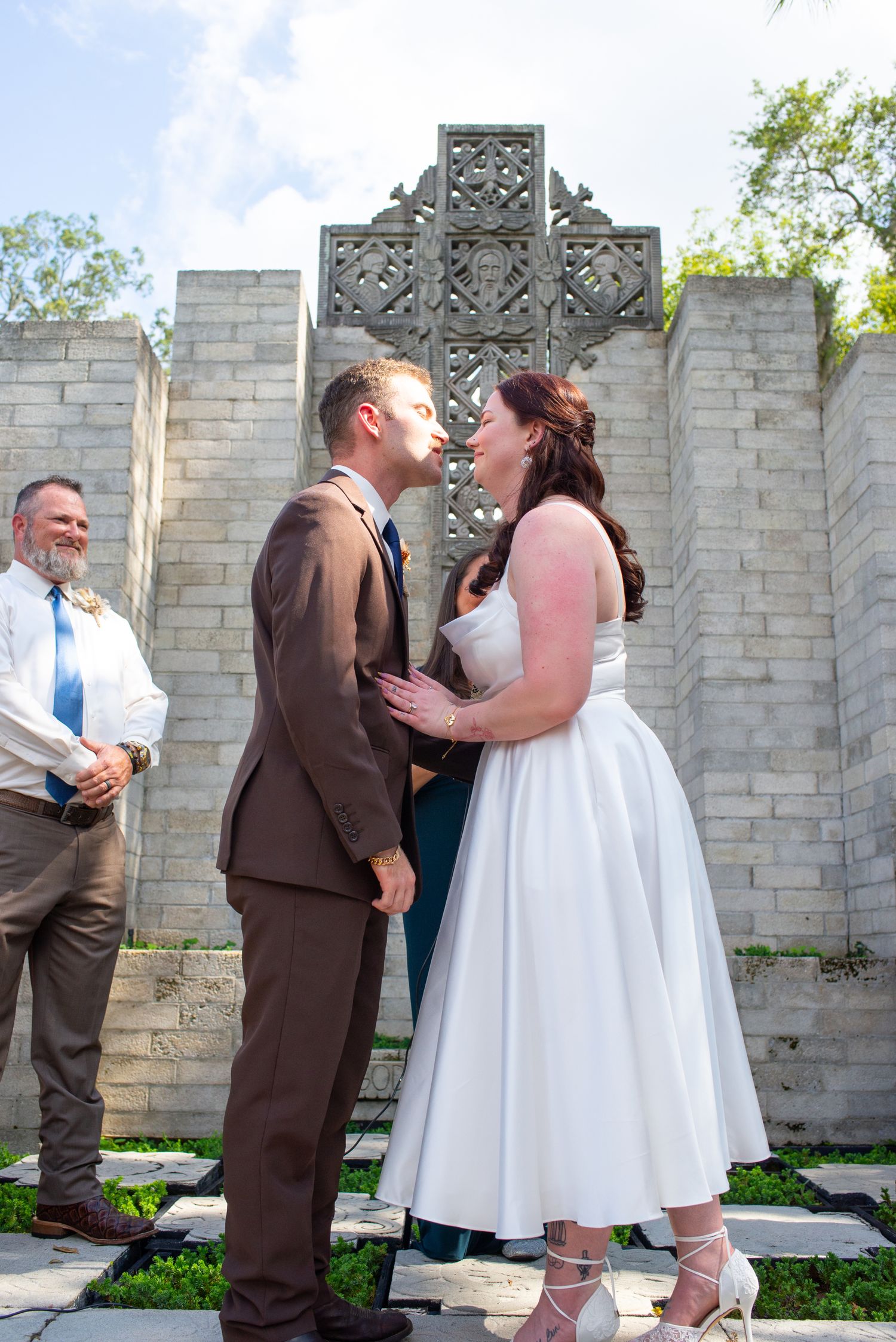 A smiling bride and groom stand face to face before a decorative stone cross during their ceremony.