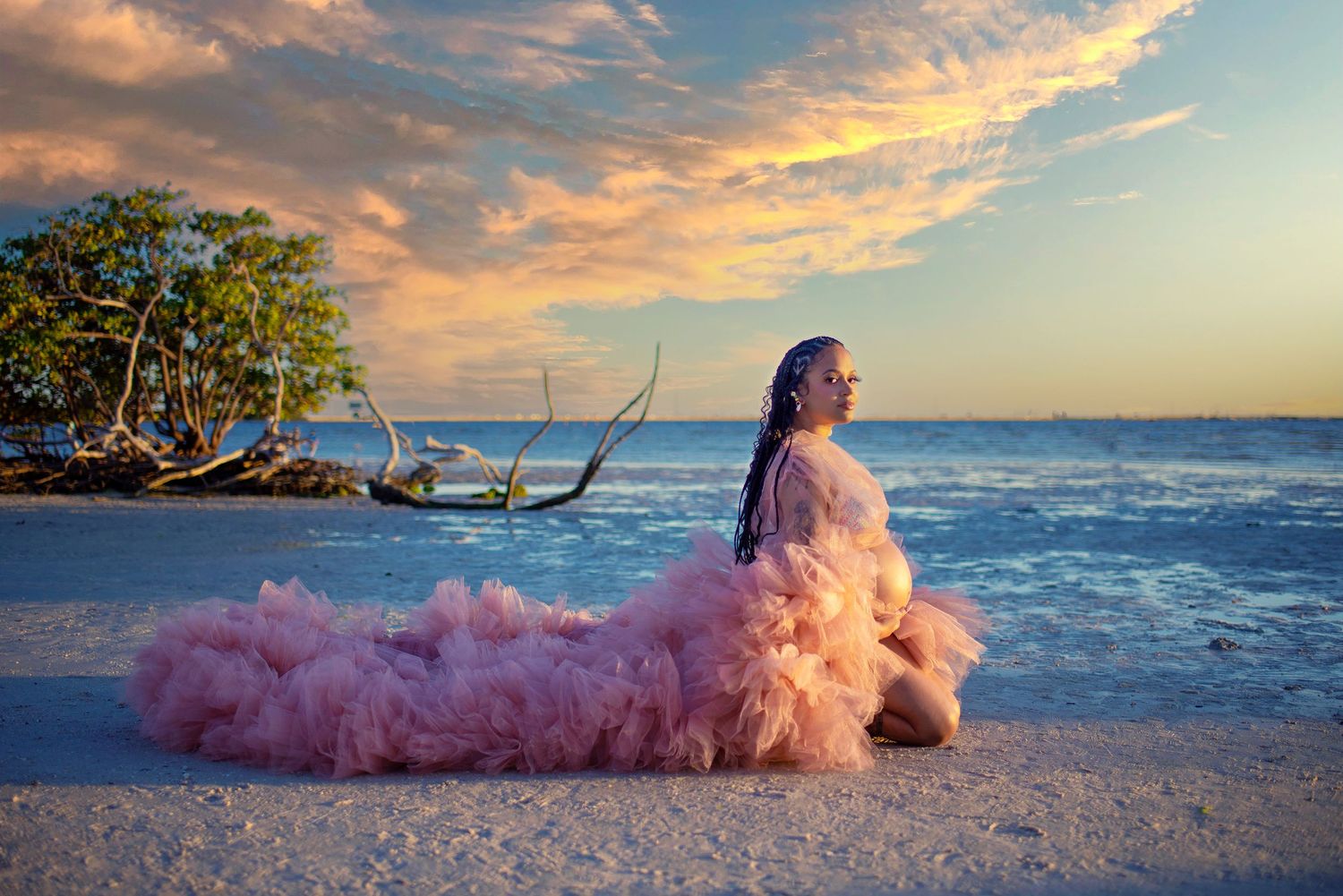 Pregnant woman in pink tulle gown kneeling on white sand beach at vibrant sunset.