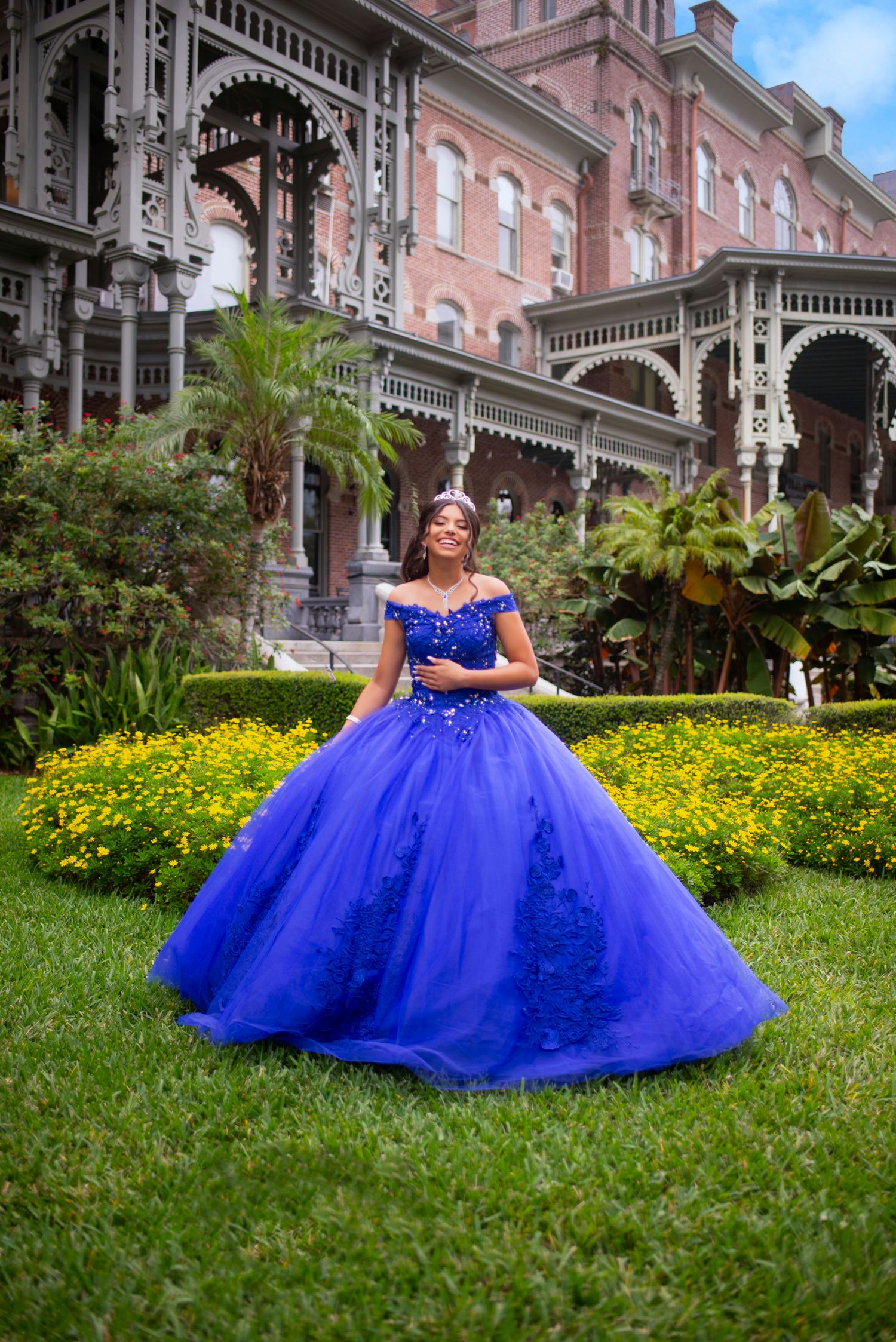 Young woman in royal blue quinceañera ball gown with tiara posing in garden of historic ornate building.