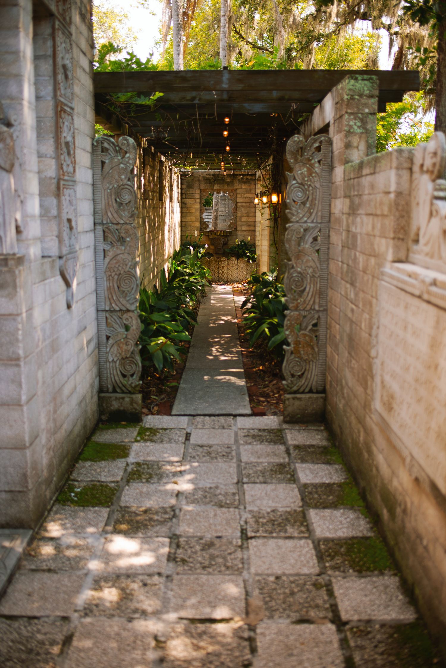Stone garden pathway with checkered tiles, ornate carvings, and lush greenery leads to an archway.