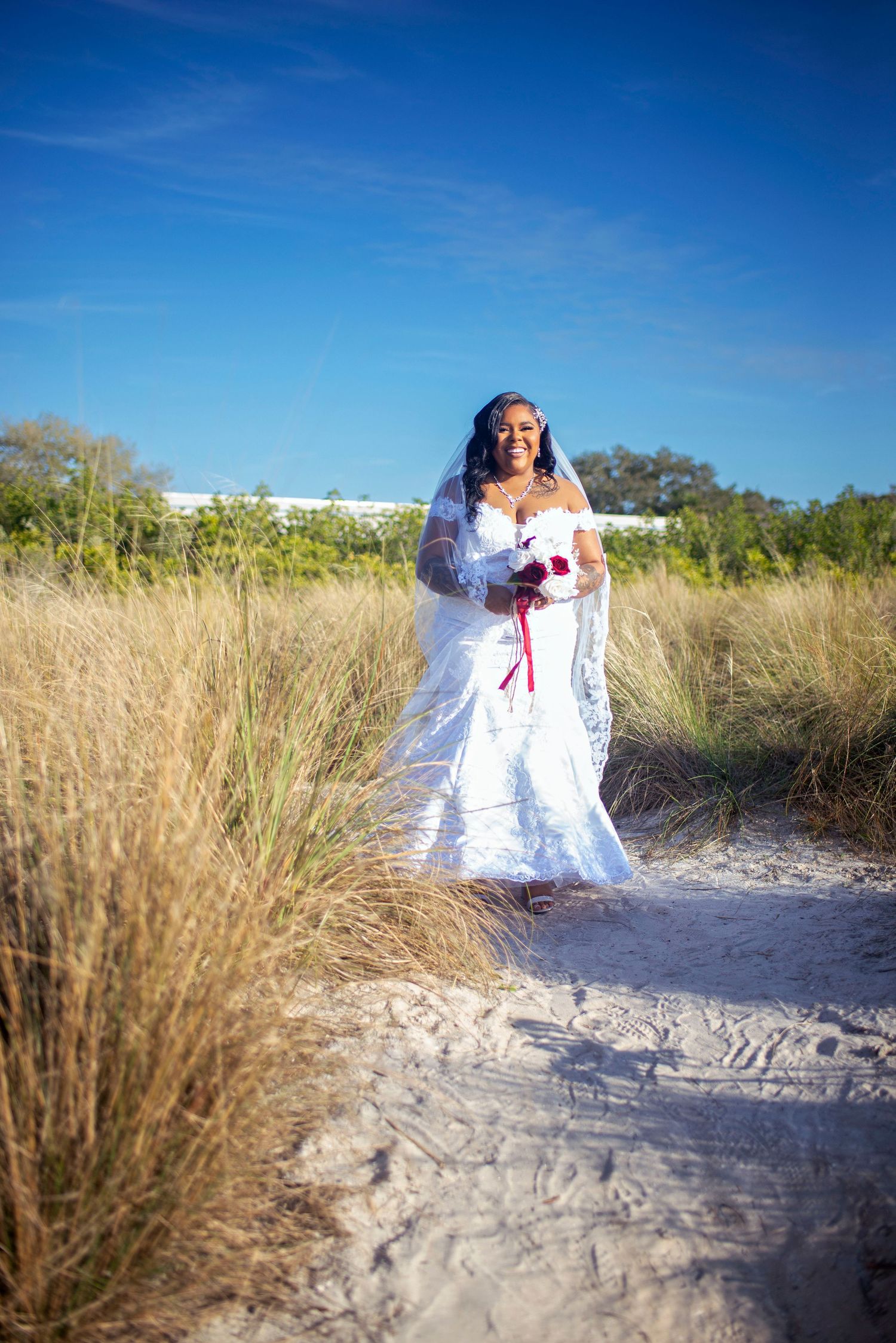 Bride in white mermaid gown with red bouquet stands on sandy beach path under blue sky.