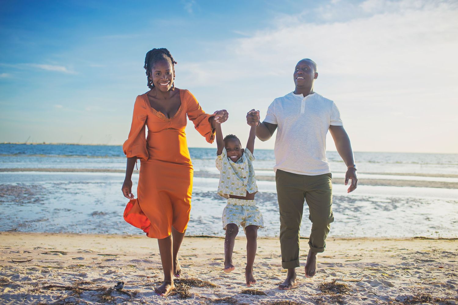 A happy family swings their child between them while walking on a sunny beach.