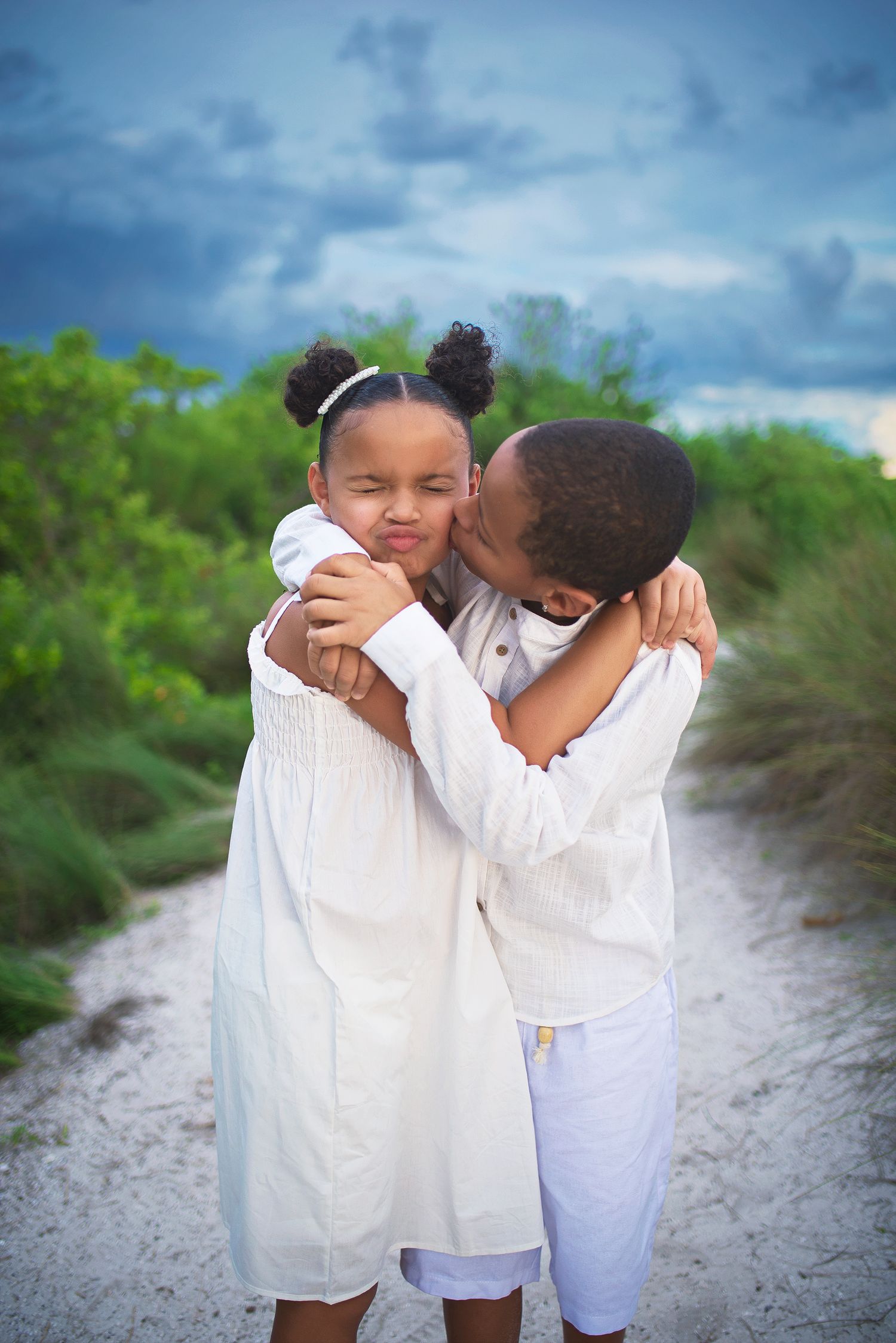 Affectionate boy in white outfit kissing his younger sister on cheek on sandy nature path.