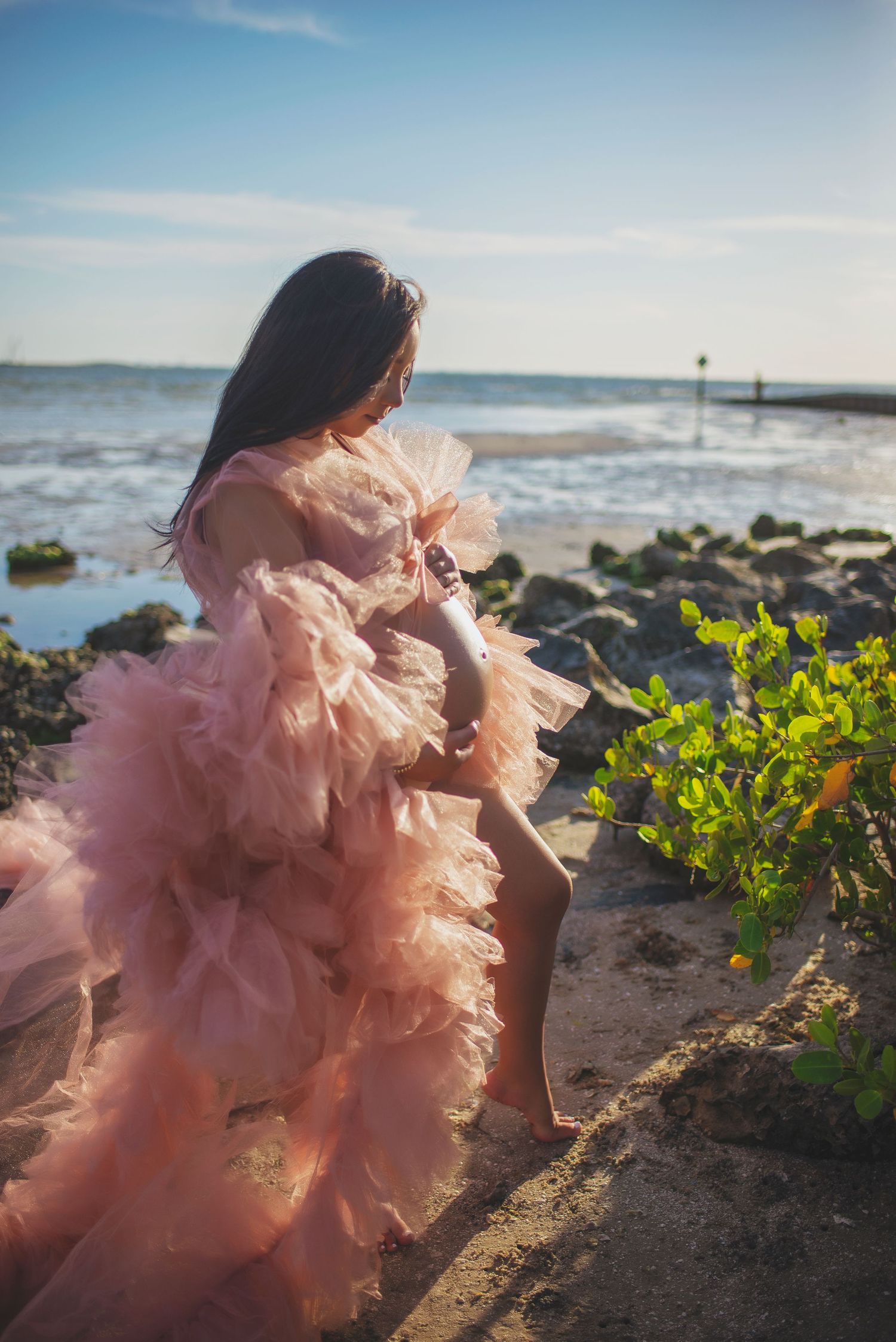 Pregnant woman in pink ruffled maternity gown sitting on rocky shoreline at golden sunset.