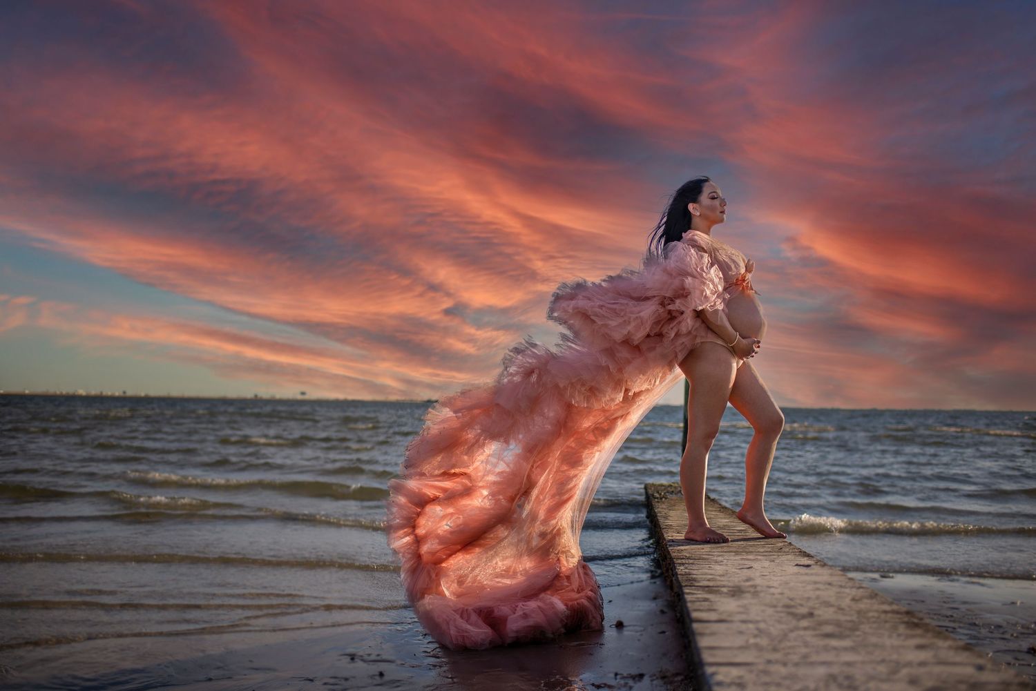 Woman in stunning pink ruffled gown standing on pier with dramatic red sunset sky over water.