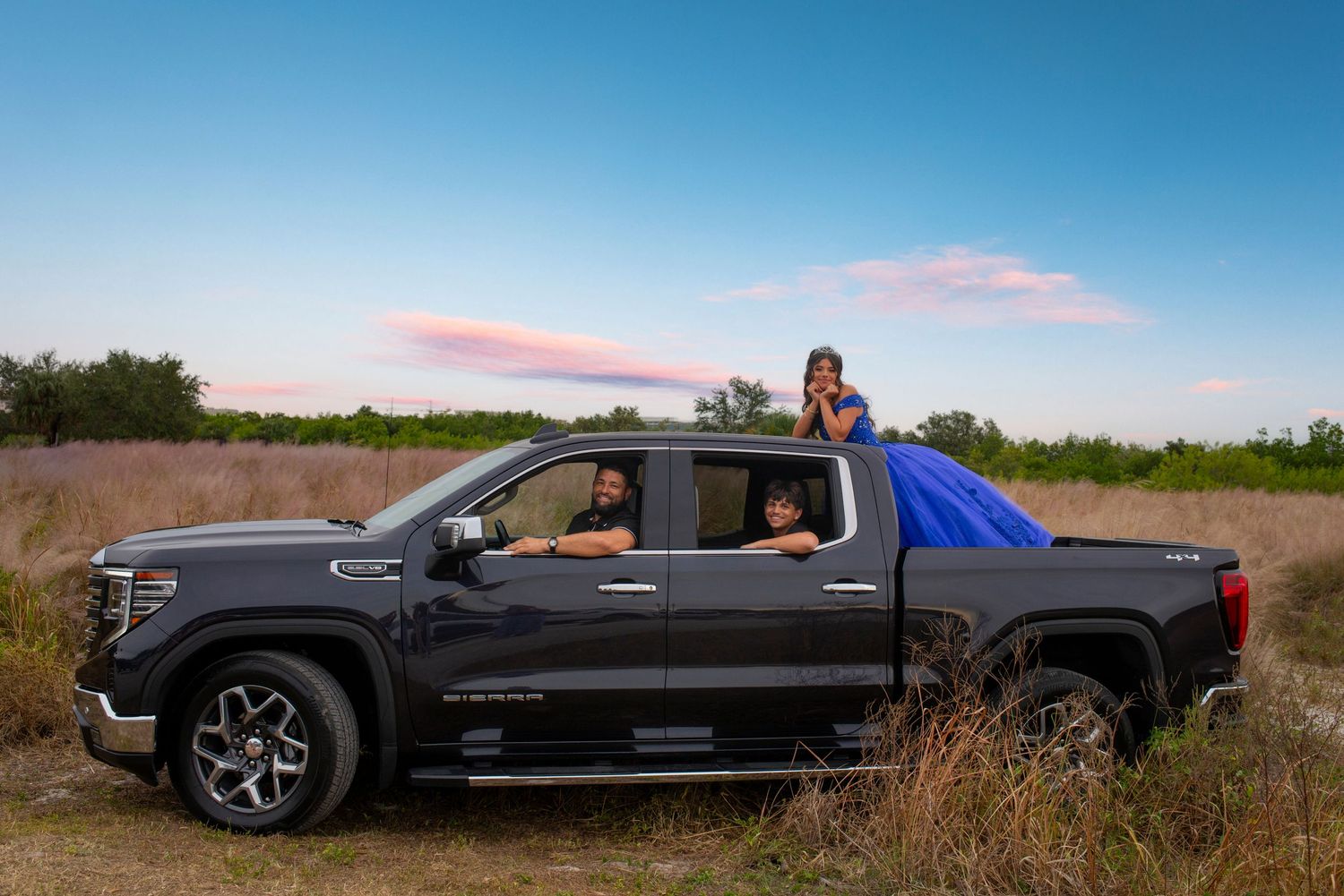 A group of people pose with a dark GMC pickup truck in a rural field at sunset.