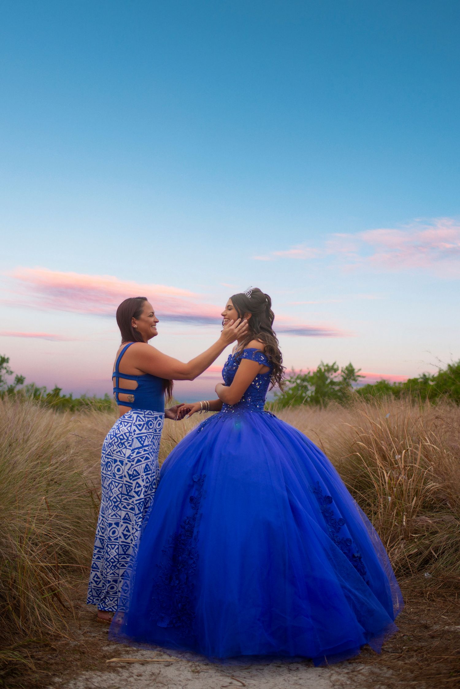Two women in matching blue dresses share a tender moment in a field at dusk.
