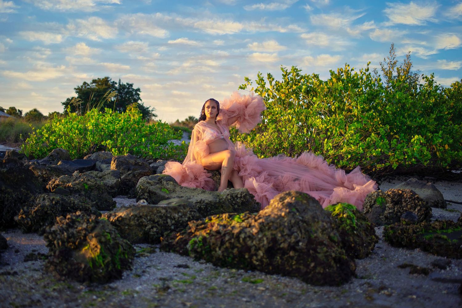 Woman in flowing pink tulle gown sitting on mossy rocks surrounded by lush greenery under a dramatic cloudy sky.