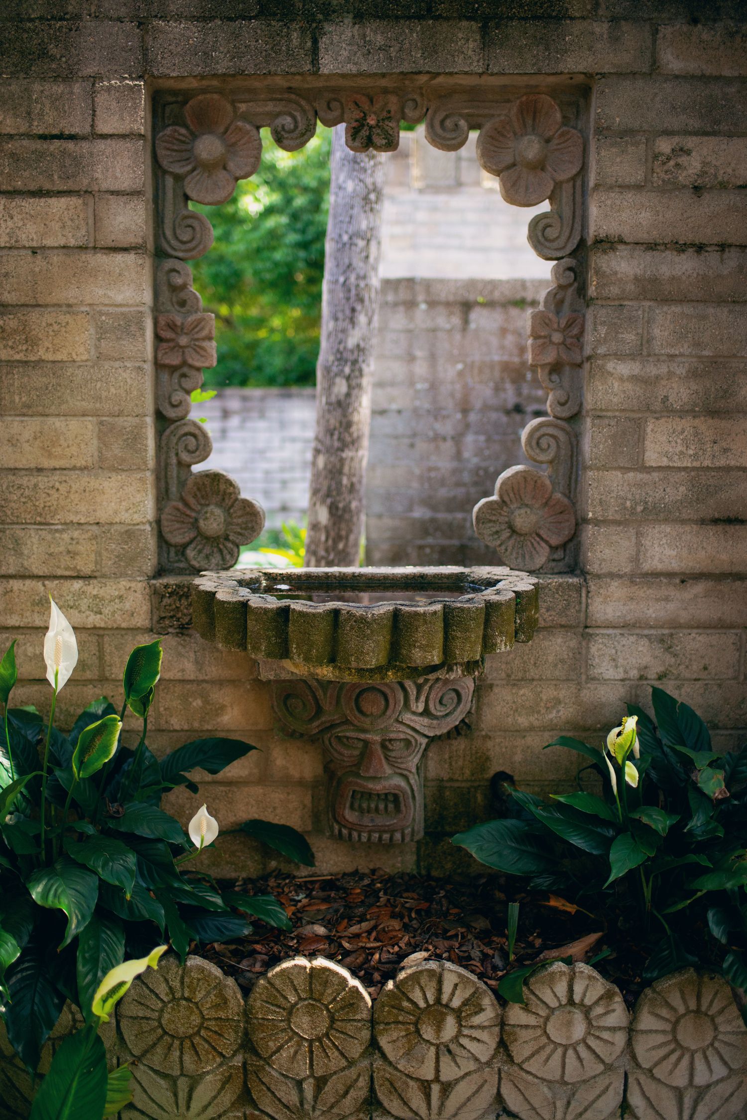 Ancient stone fountain with decorative carved frame and face motif surrounded by tropical plants in a garden wall.