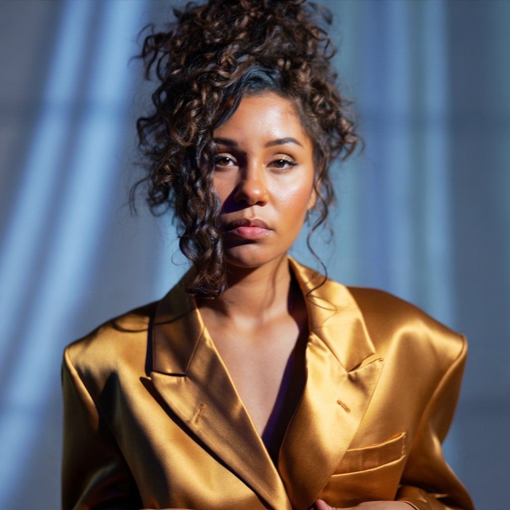 A woman with curly hair wearing a gold satin blazer poses in dramatic blue lighting.