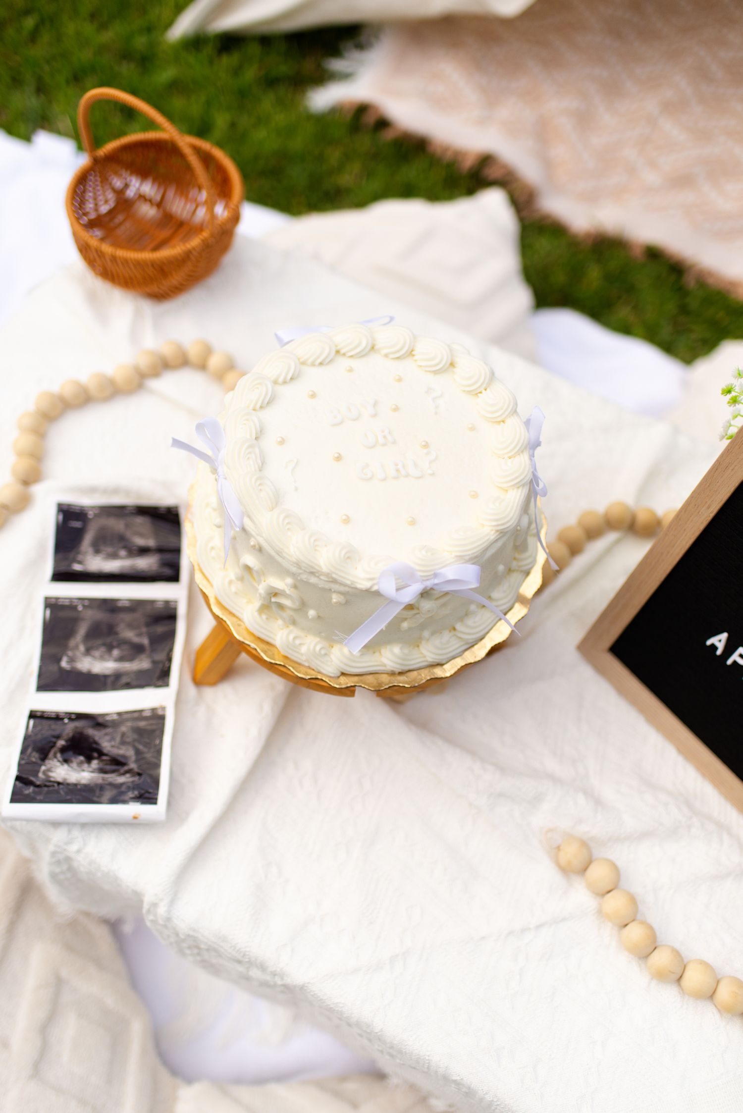 Baby shower flat lay with white cake, ultrasound photos, wooden beads, and letter board on white blanket.