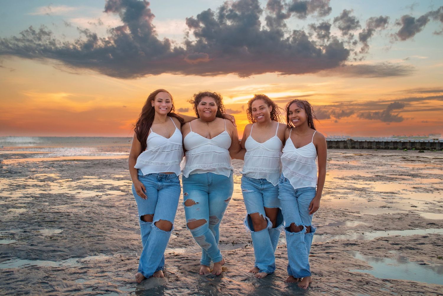 Four young women in white tops and ripped jeans pose together on a beach at sunset.