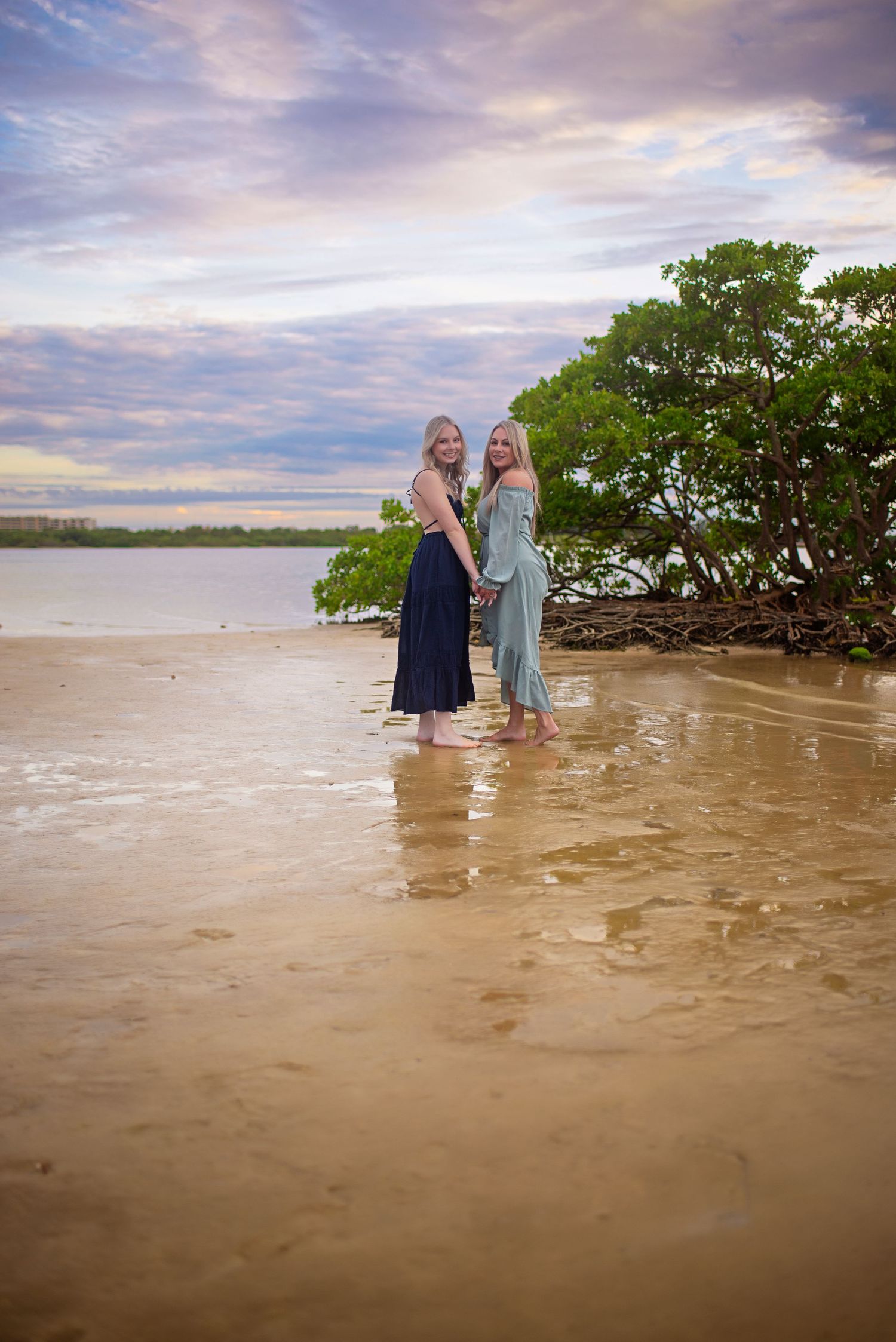 Two blonde women in flowing dresses holding hands while standing in shallow water at sunset beach.