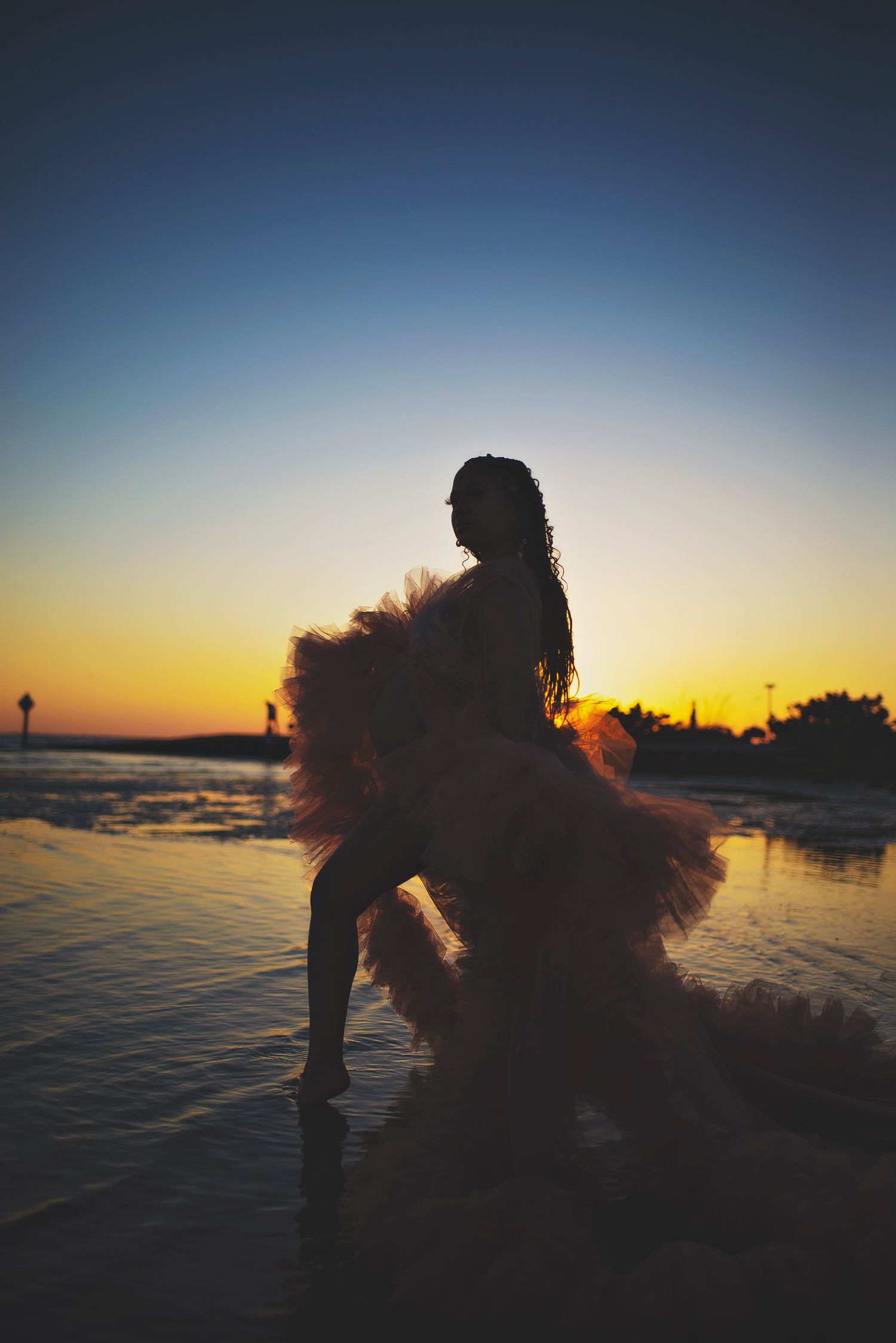 Silhouette of a woman in a ruffled tulle gown standing in shallow water during a vibrant golden sunset.