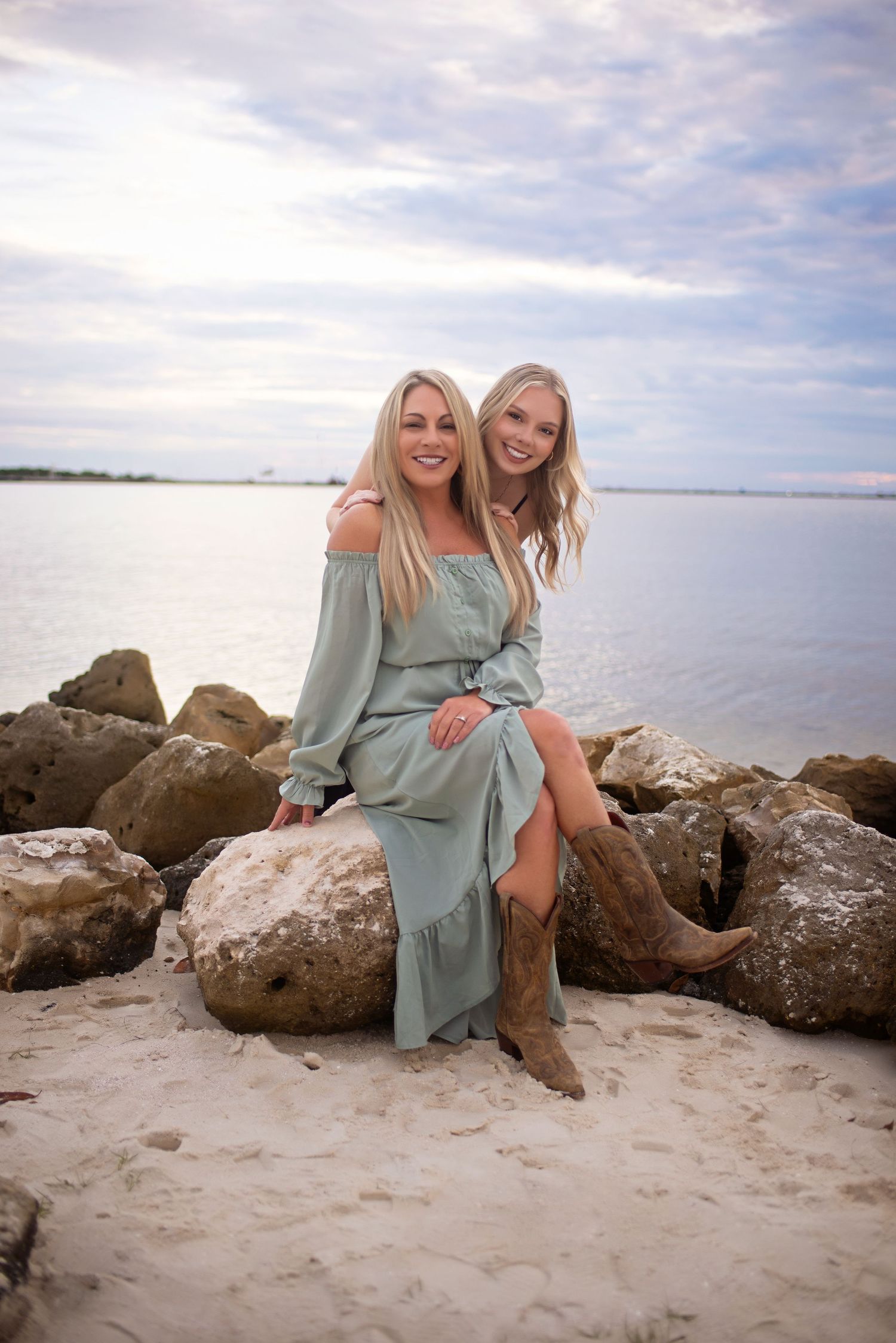Two blonde women in sage green dresses sitting together on coastal rocks with calm water in background.