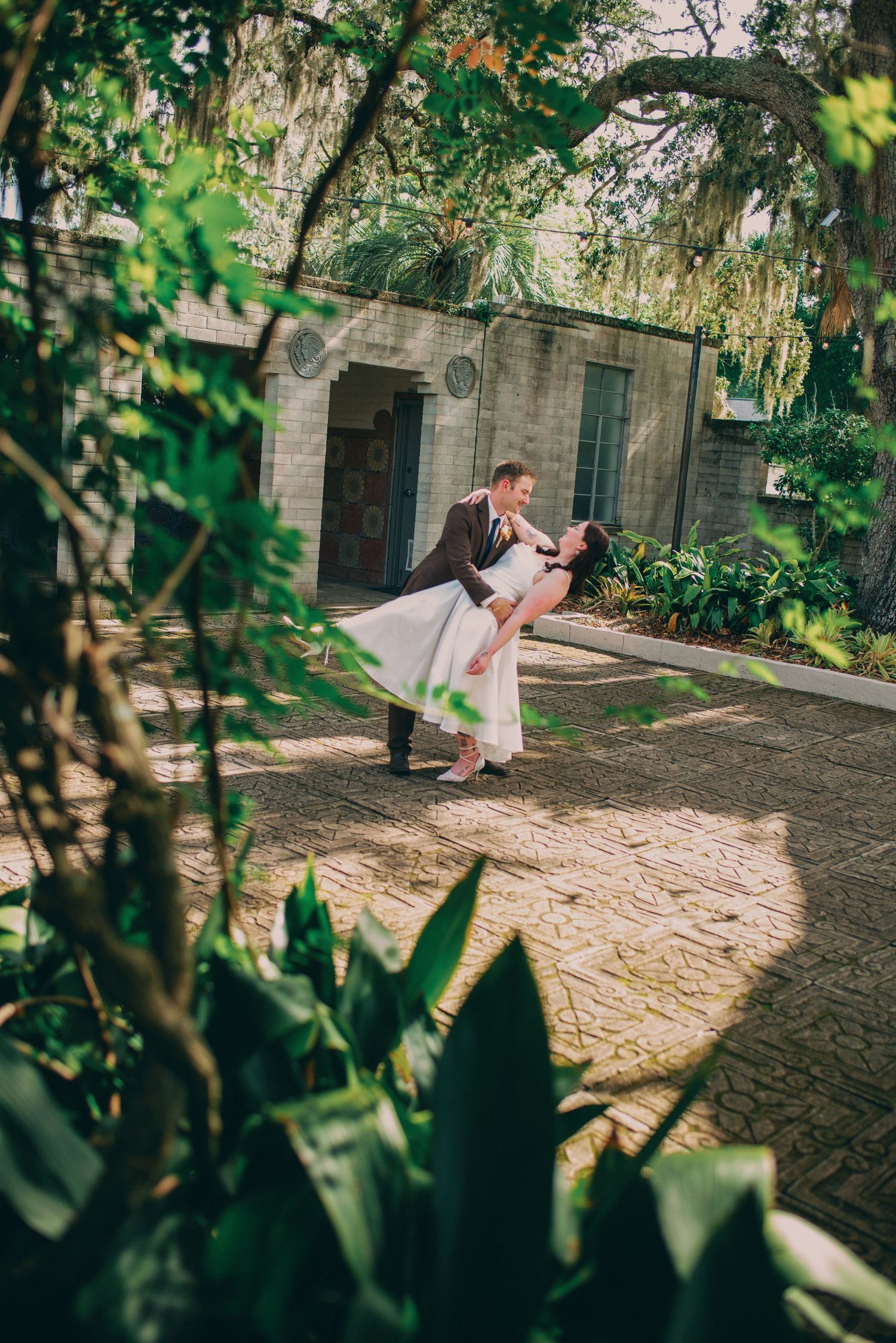 Groom dipping bride in romantic dance pose outdoors at lush garden wedding venue with stone architecture.
