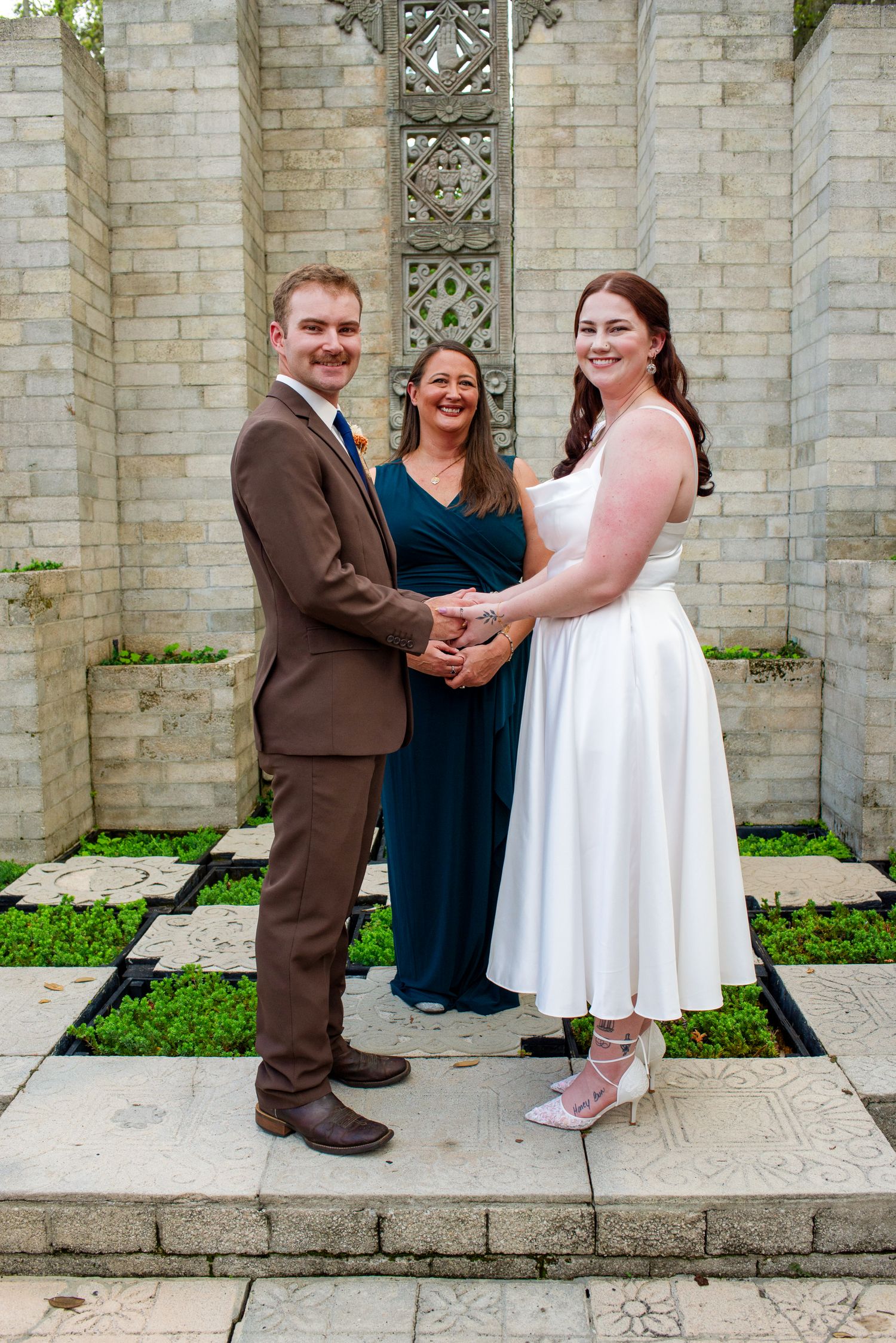 Bride in tea-length white dress and groom in brown suit hold hands with officiant at outdoor wedding.