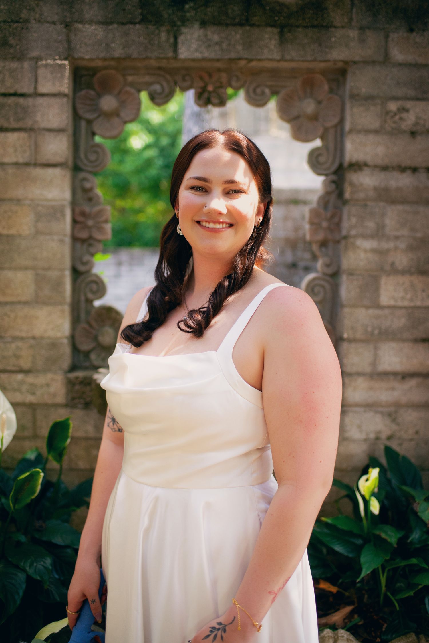 Smiling bride in white spaghetti strap dress poses before decorative carved stone wall outdoors.