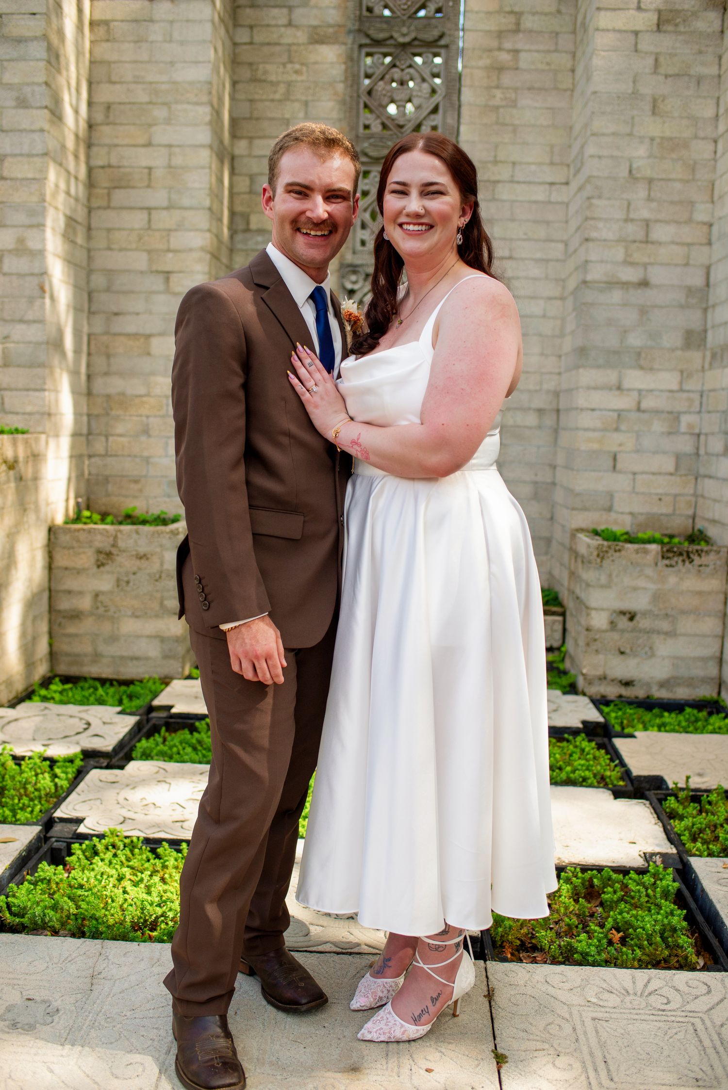 A happy bride and groom pose together in front of an ornate stone wall after their wedding.