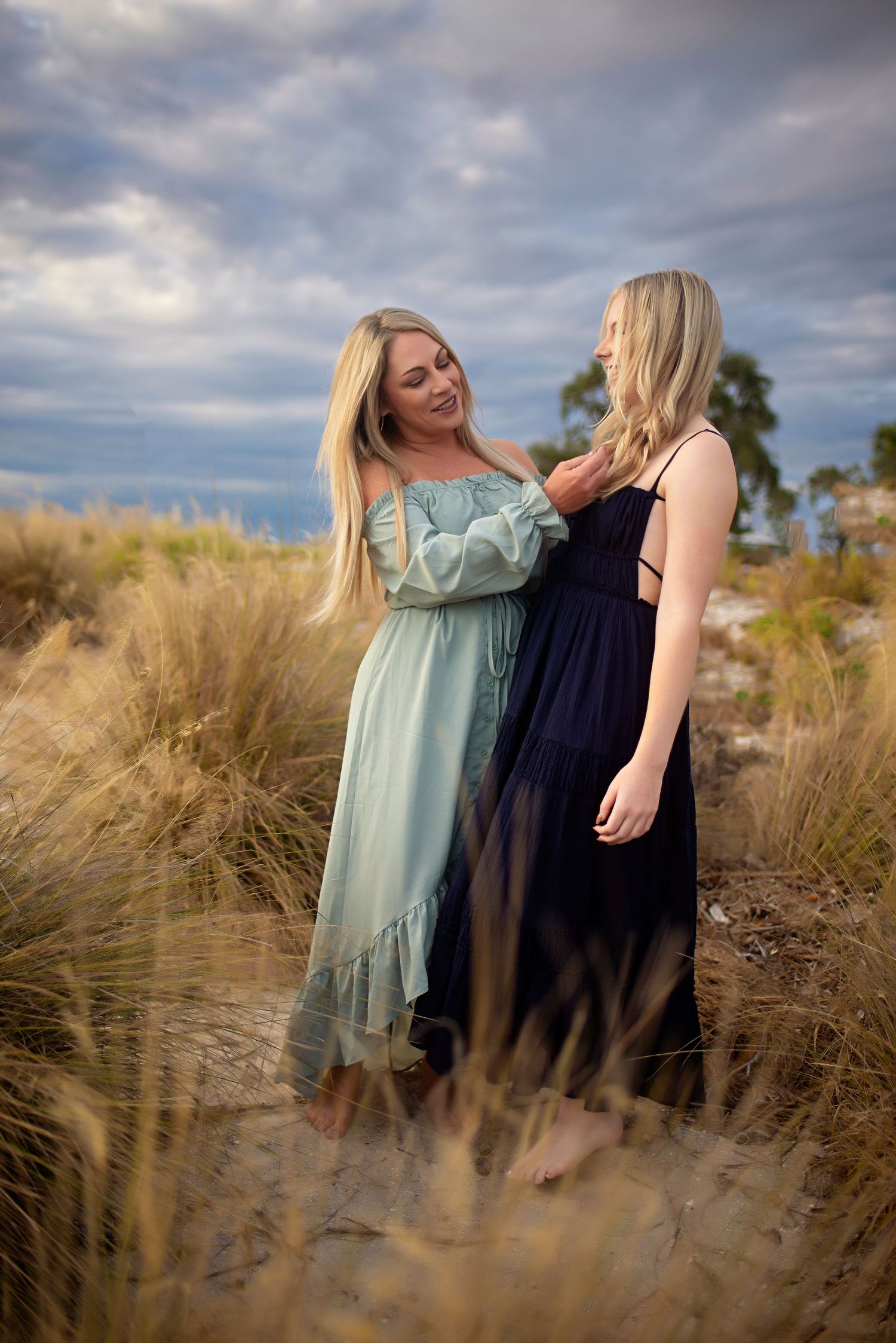 Two blonde women in elegant dresses share a tender moment on a sandy beach.