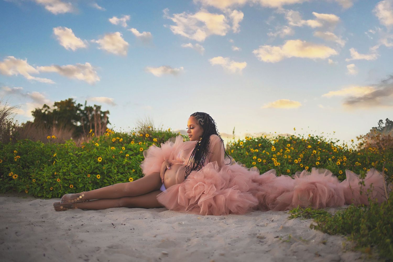 Woman in pink tulle ruffled gown sitting gracefully on sandy ground surrounded by yellow wildflowers at sunset.