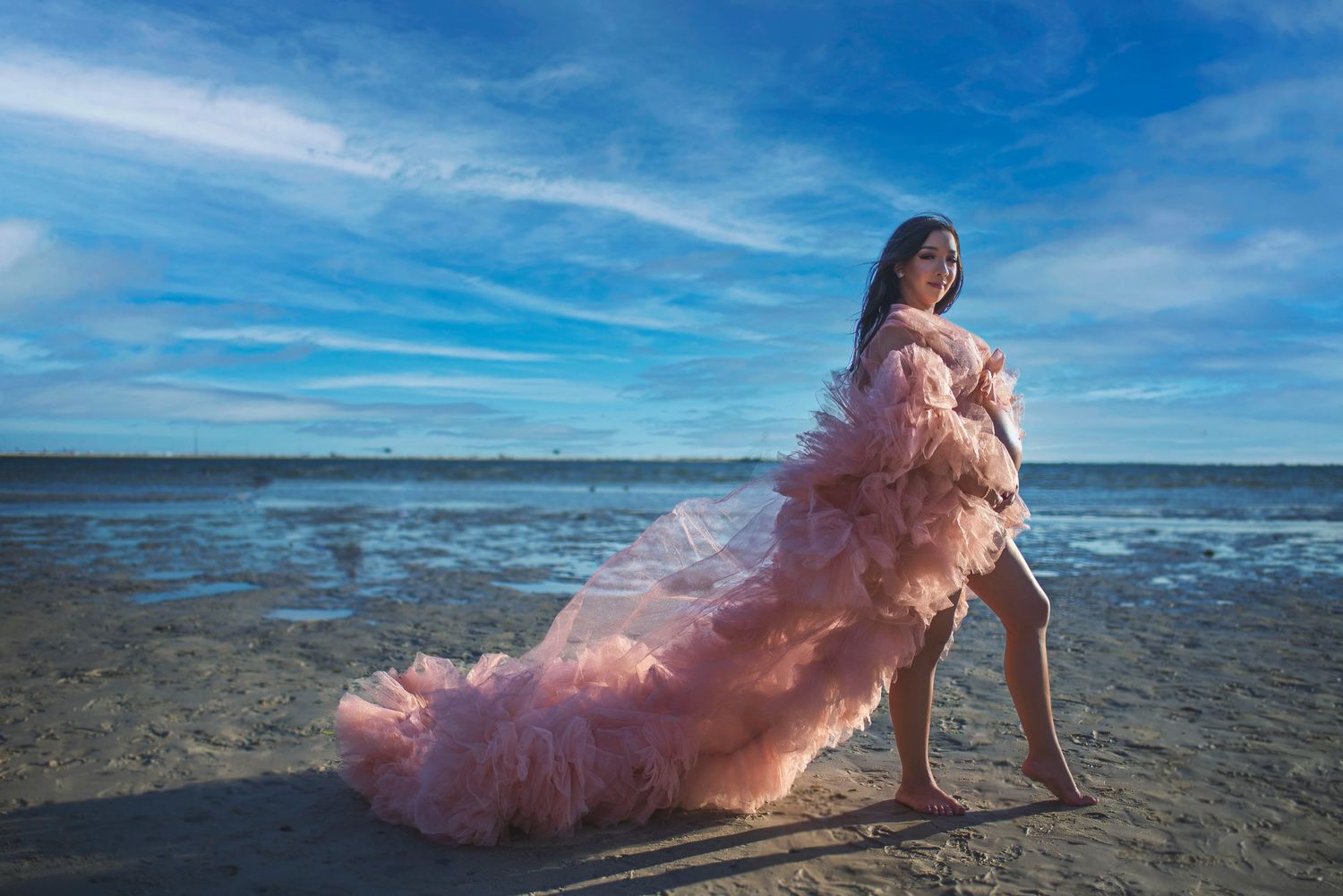 Pregnant woman in elegant pink tulle maternity gown posing on sandy beach with ocean backdrop.
