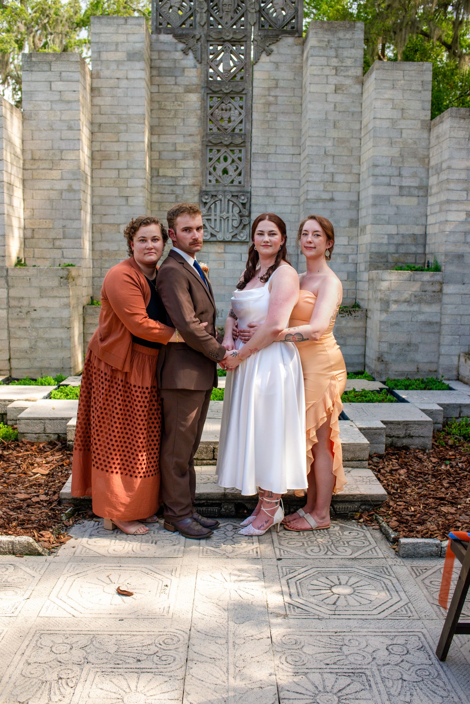 Wedding party of four poses together before ornate stone cross and brick wall at outdoor ceremony venue.