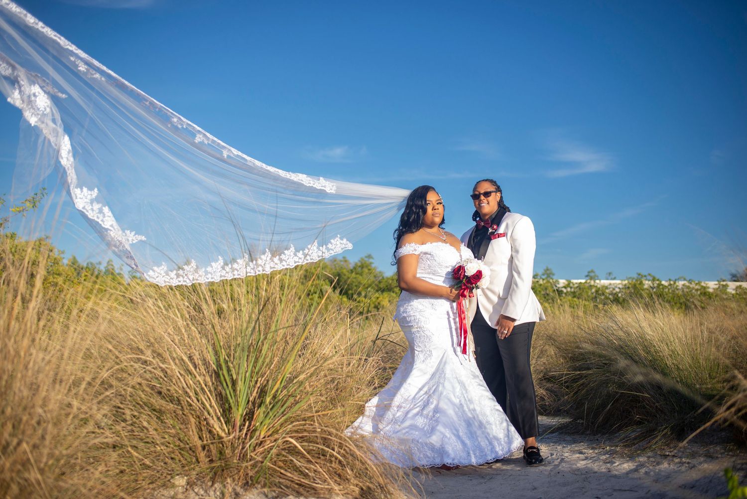 Wedding couple stands on white sand beach as bride's long lace veil billows in the wind.
