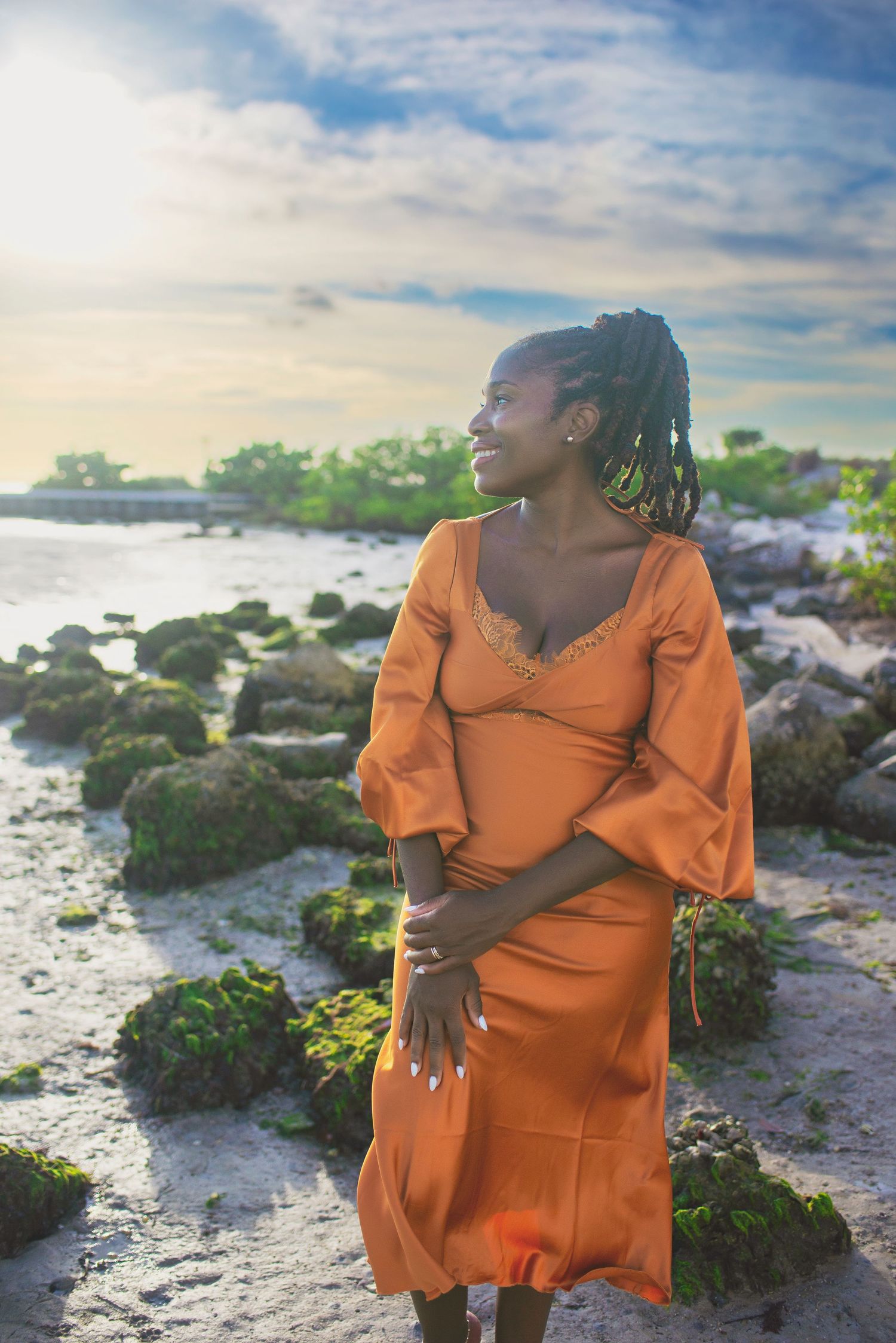 A woman in an orange dress stands on a rocky shoreline at golden hour sunset.