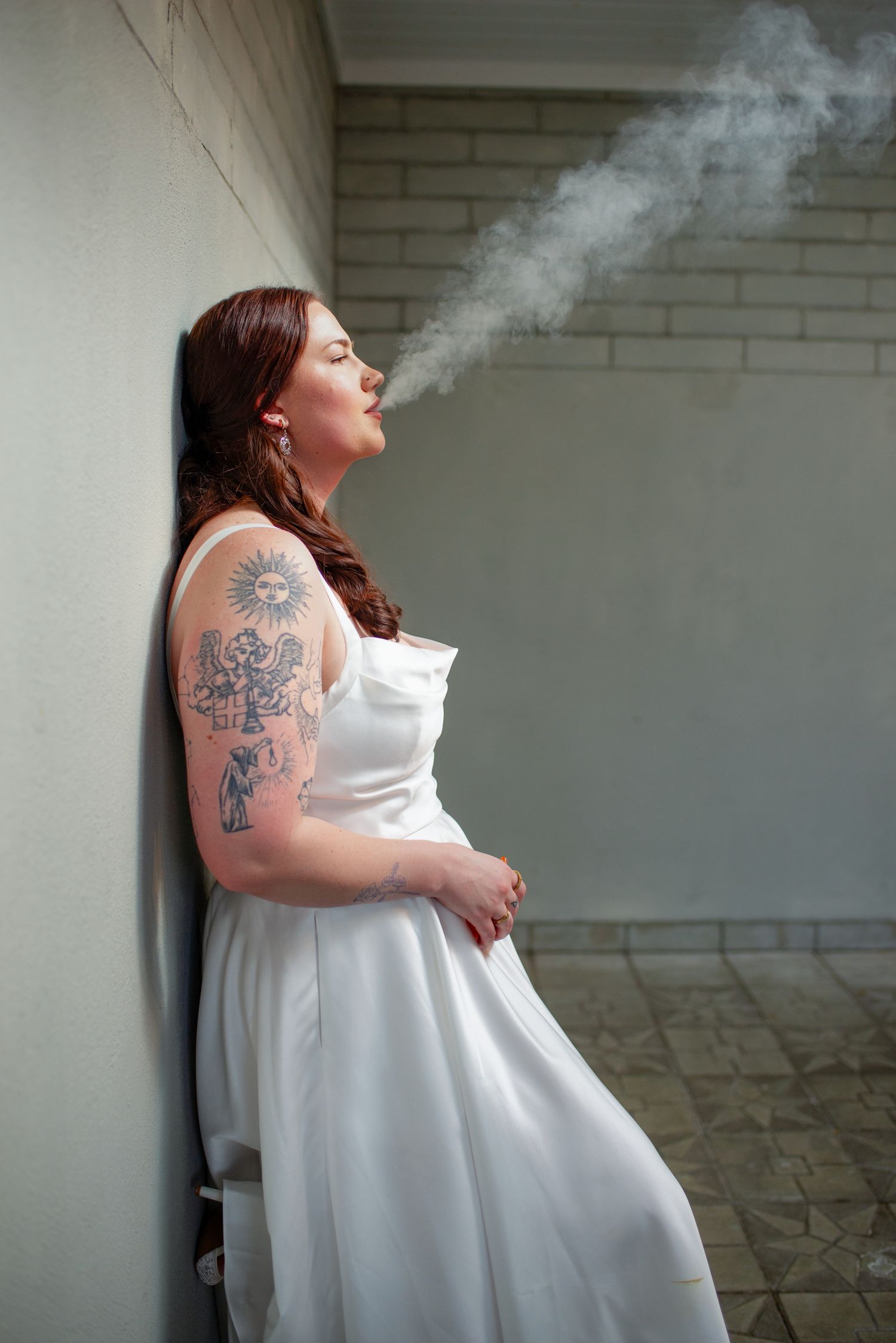 Tattooed bride in white satin dress exhales smoke while leaning against a wall before her wedding ceremony.
