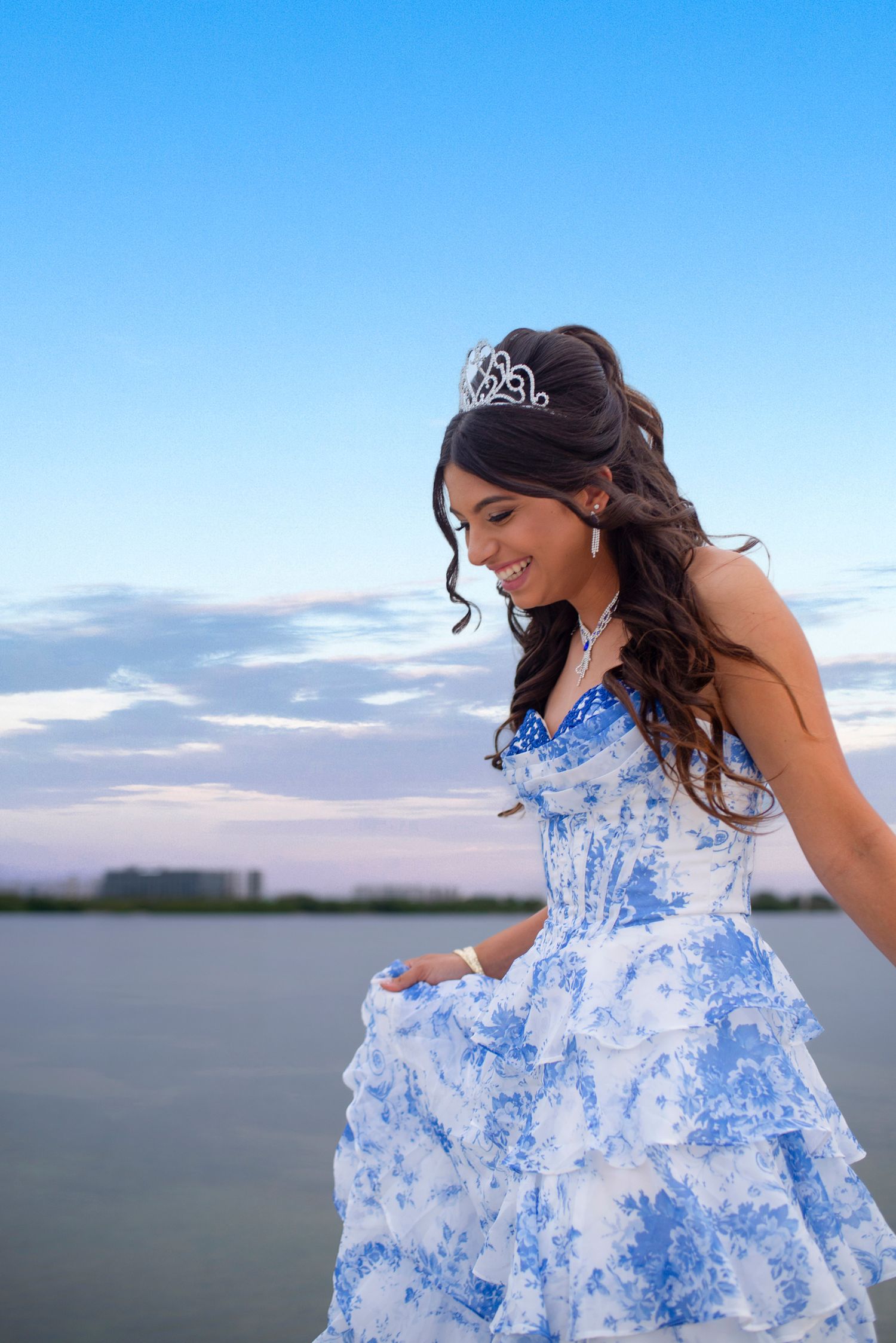 A young woman in a blue and white floral quinceañera gown smiles wearing a tiara by the water.