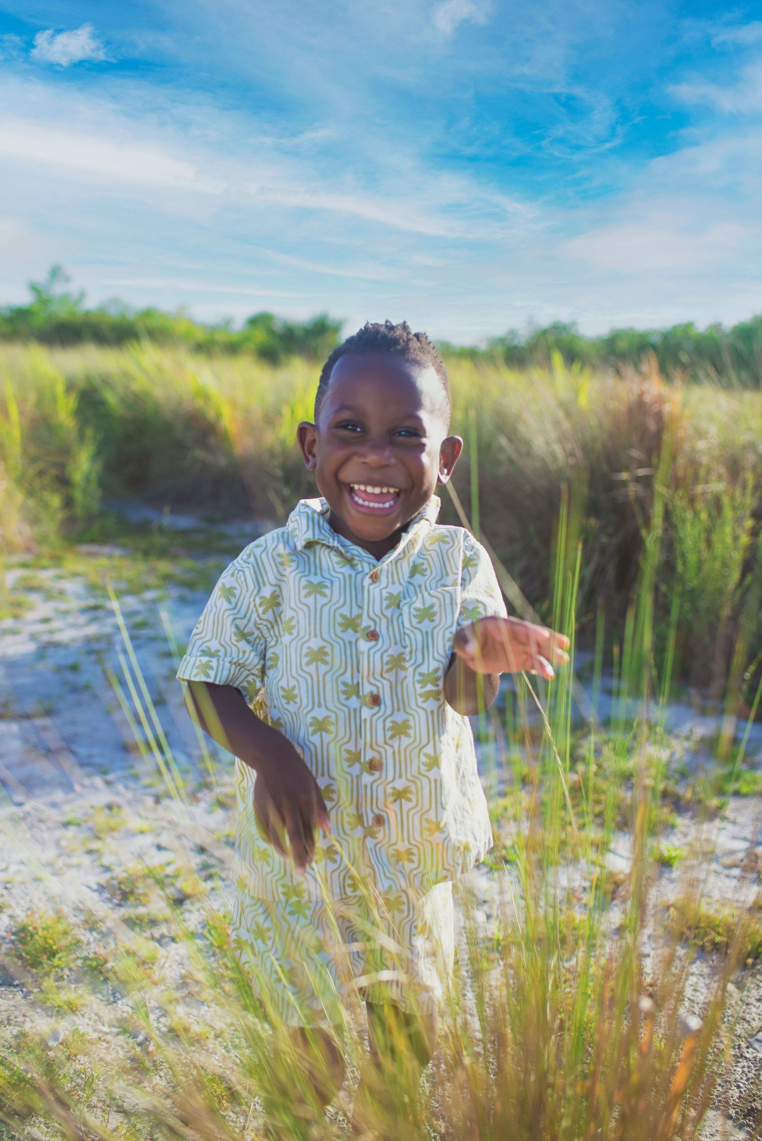 A smiling young boy in a patterned shirt stands among tall grass on a sunny day.
