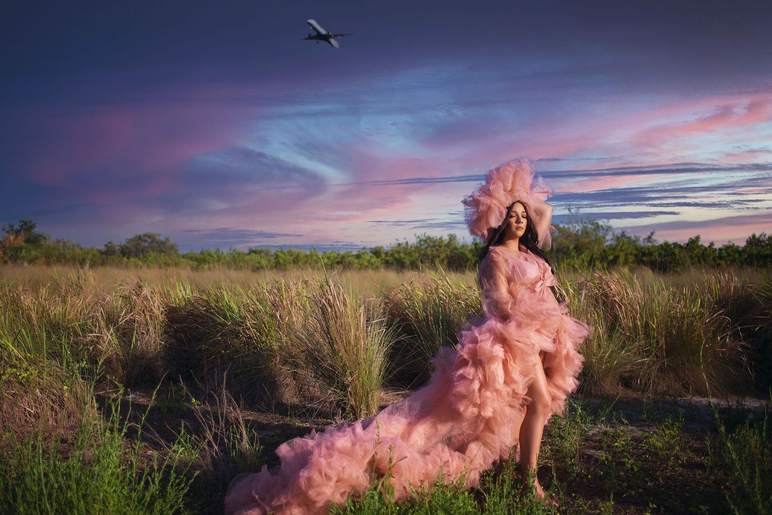 Pregnant woman in pink ruffled tulle gown posing in golden field under vibrant purple sunset sky.