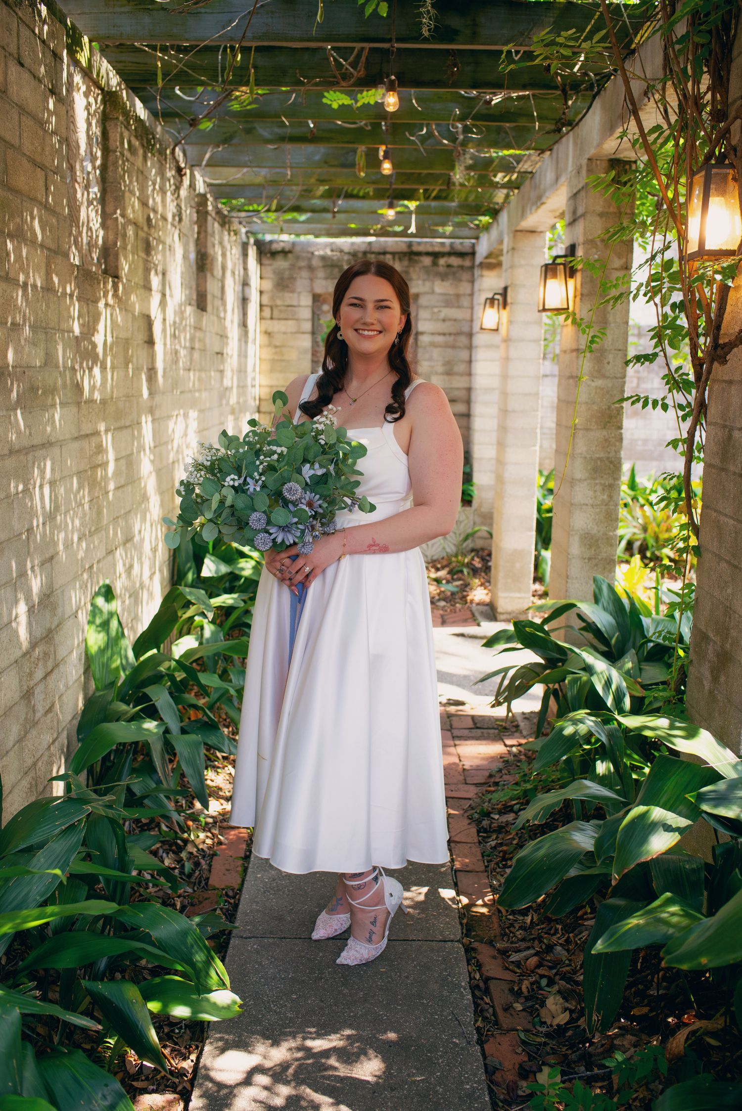 Bride in white midi dress holding lush bouquet standing in sunlit garden pergola walkway.