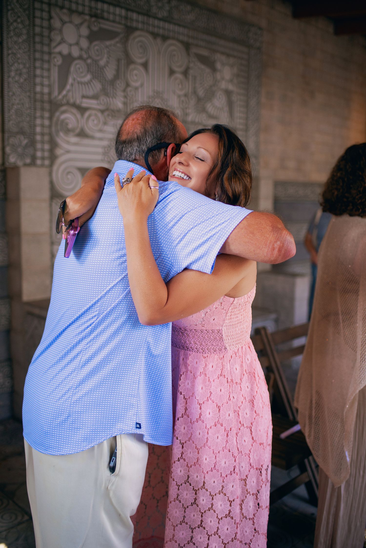 Smiling woman in pink lace dress joyfully hugs a guest at an outdoor wedding celebration.