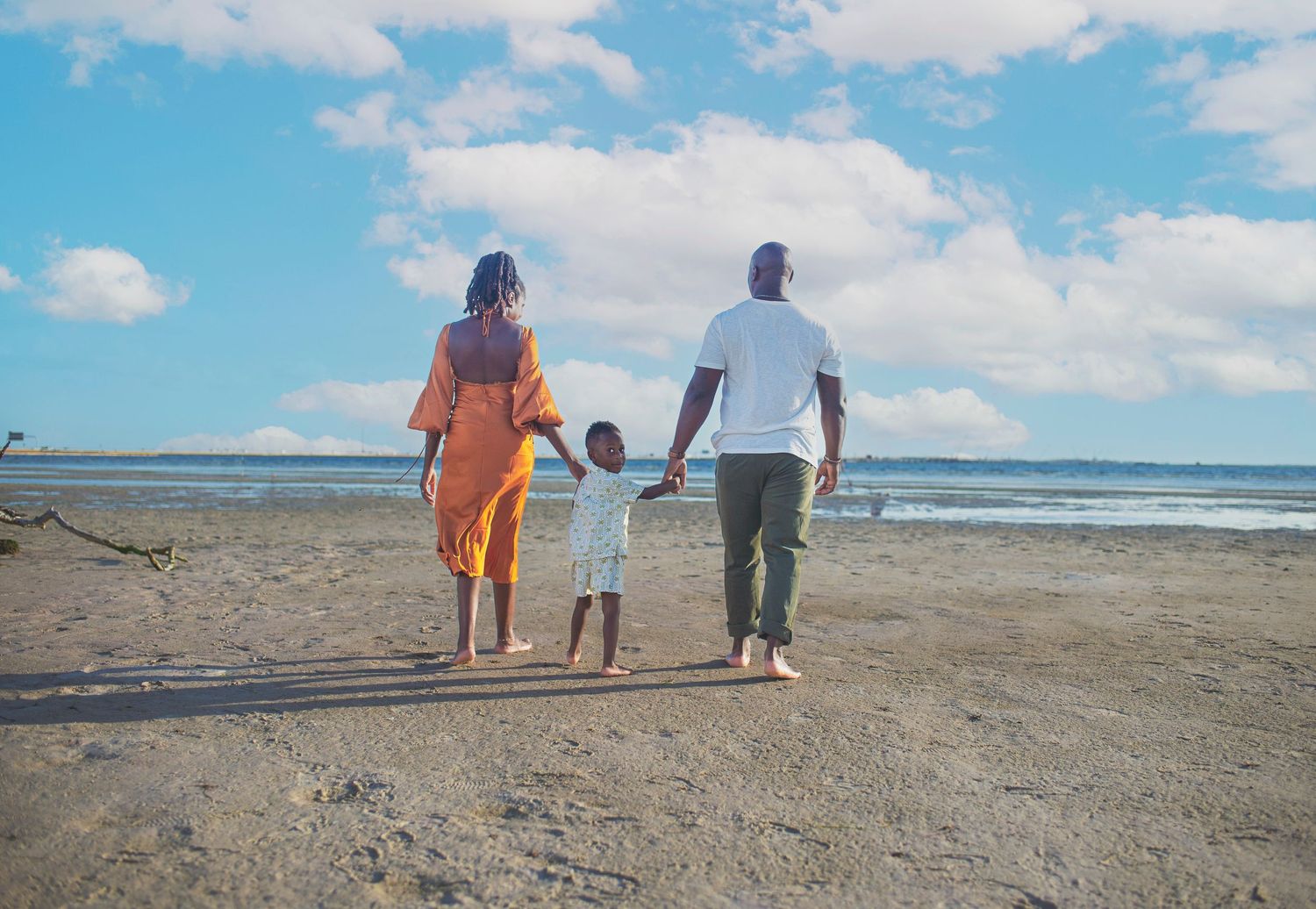 A family of three walks hand-in-hand along a sunny beach shoreline.
