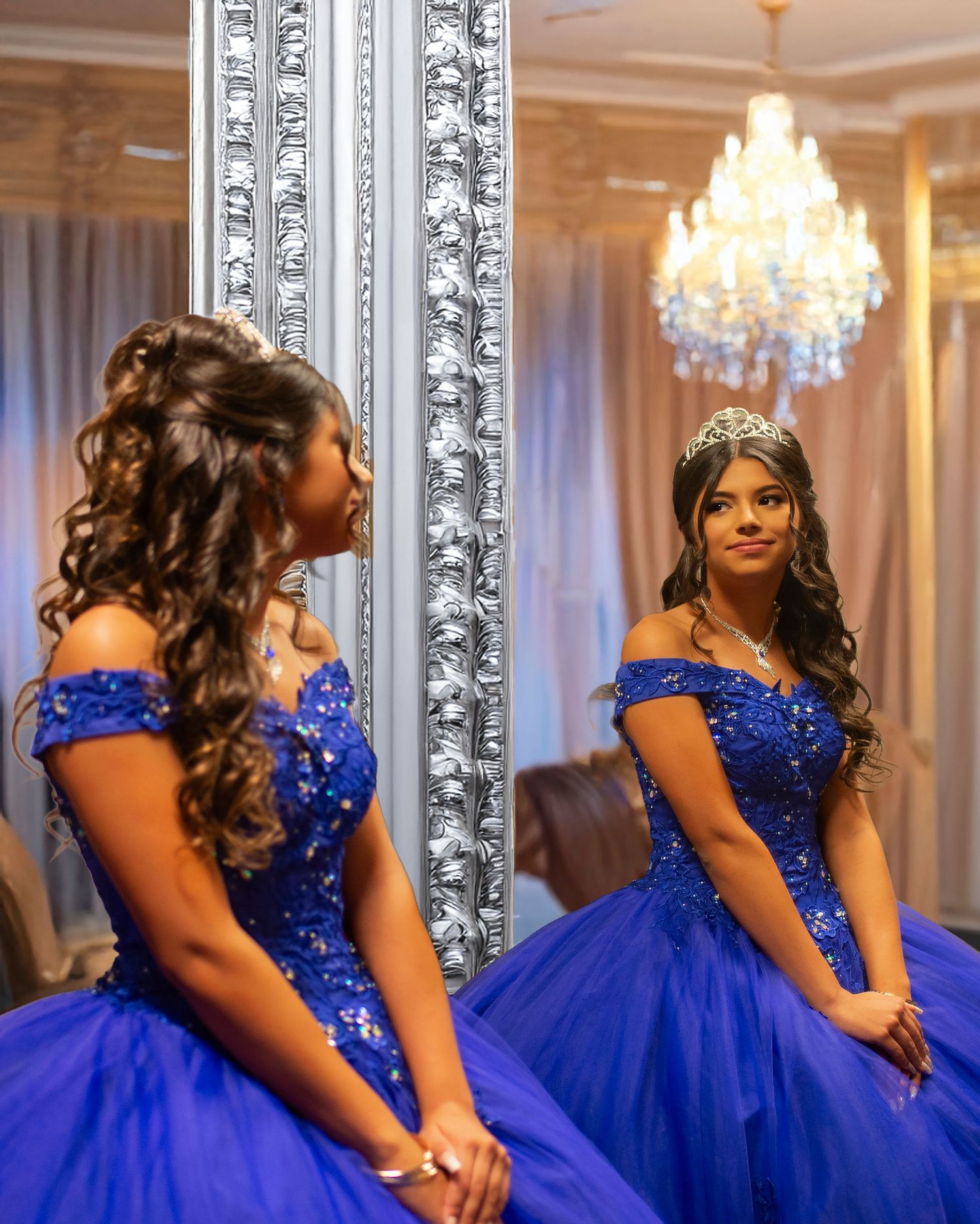 Young woman in royal blue quinceañera gown and tiara reflected in an ornate mirror under a crystal chandelier.