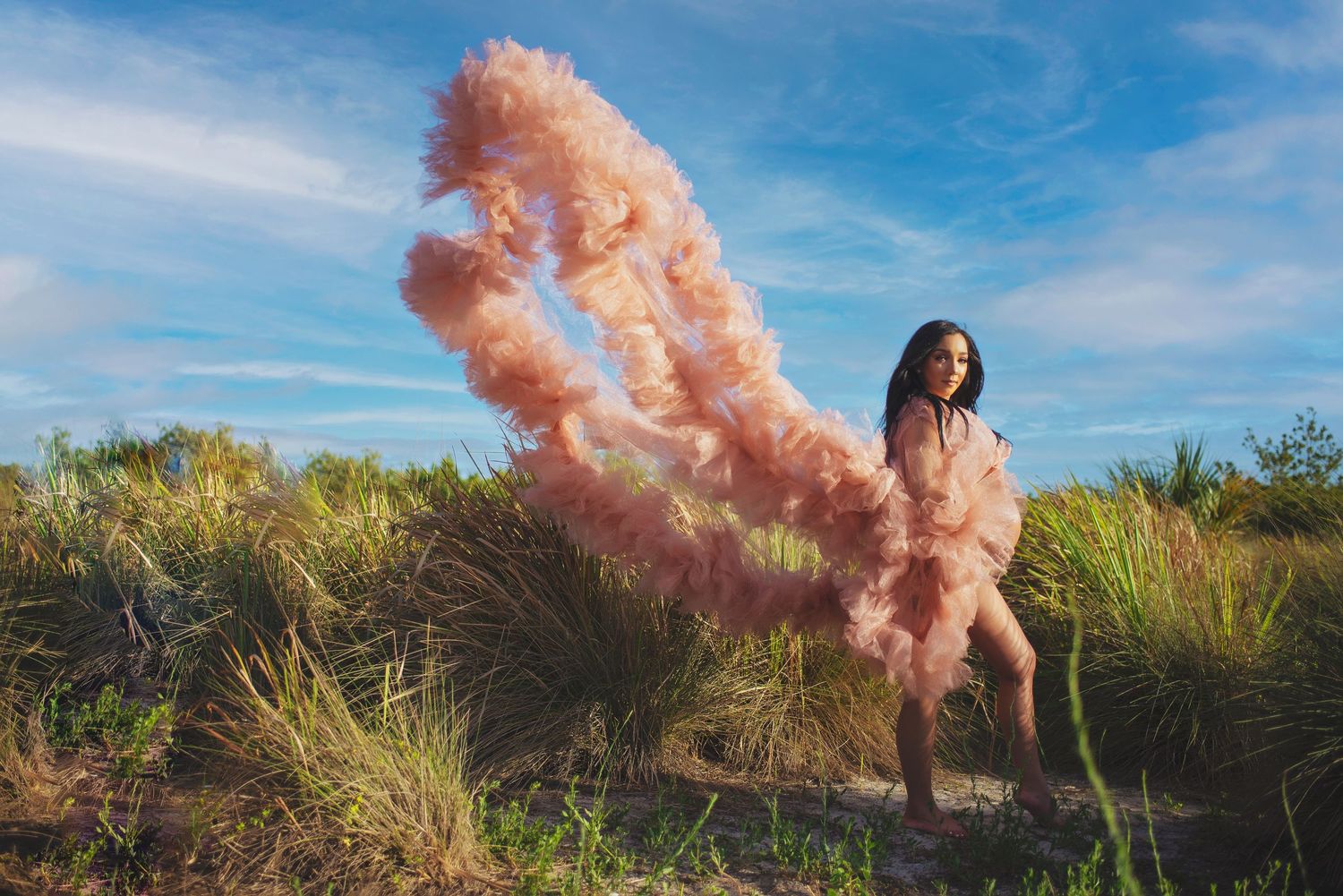 Woman in voluminous pink tulle gown with dramatic train blowing in wind on sandy dune landscape.