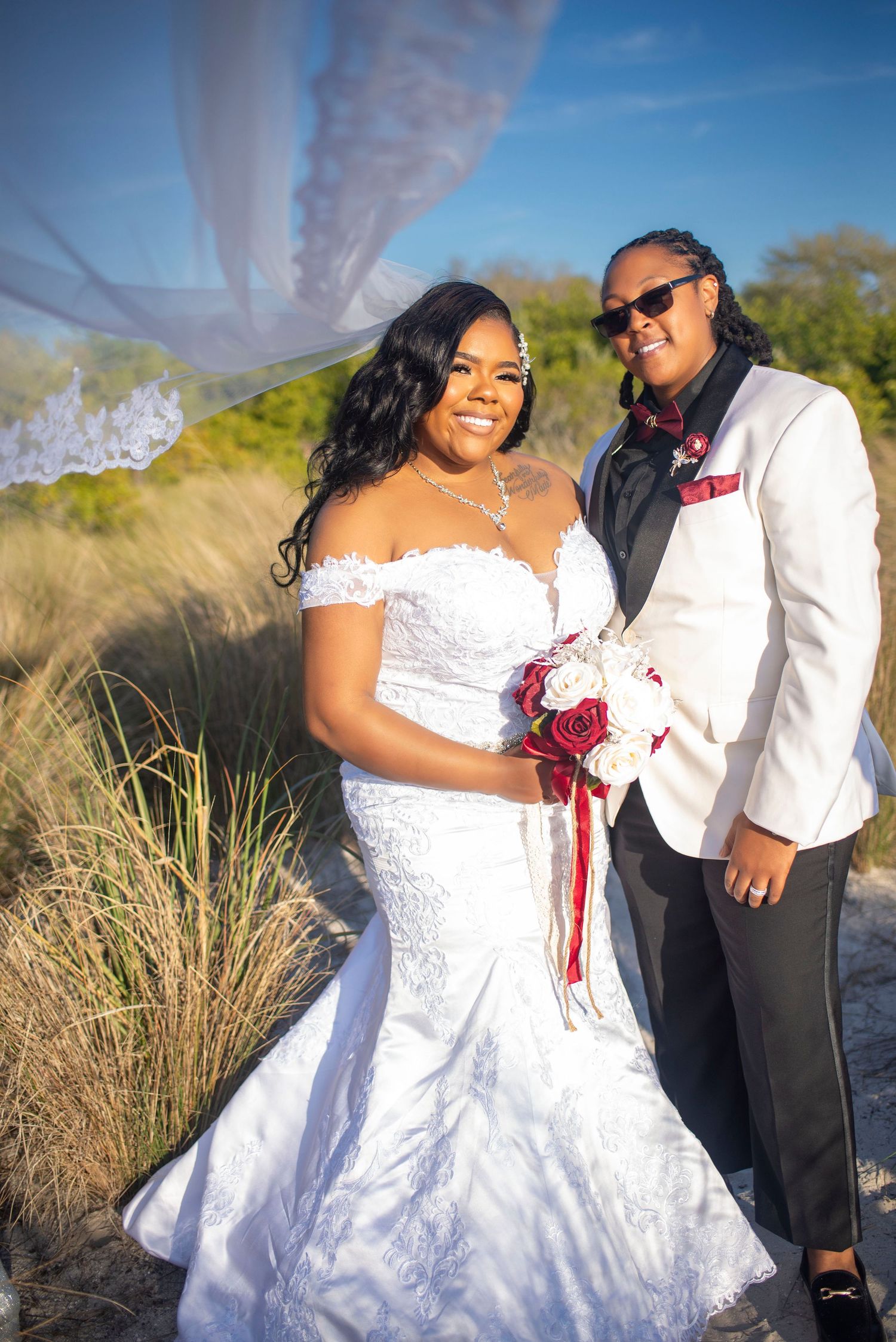 Bride in white lace mermaid gown and partner in white tuxedo pose on sandy beach dunes.