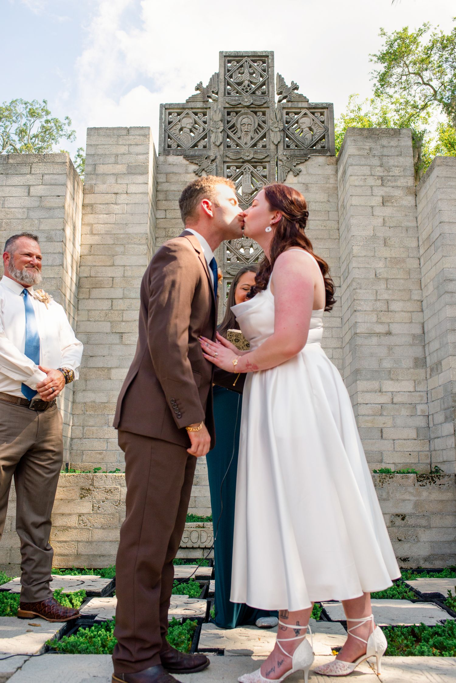 Bride and groom share first kiss at outdoor wedding ceremony before ornate stone cross and brick wall.