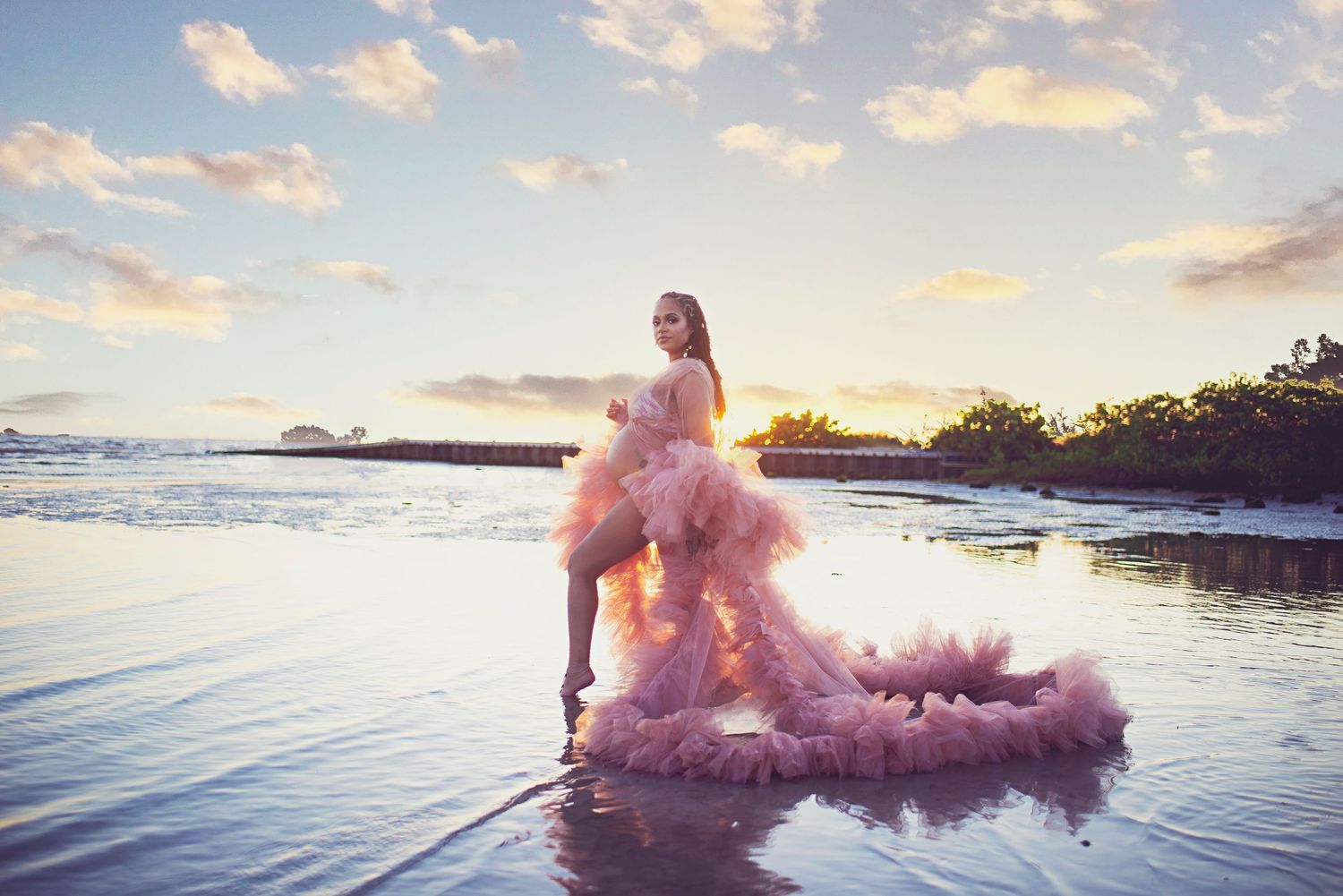 Woman in dramatic pink tulle gown posing elegantly on a reflective beach shoreline at golden sunset.