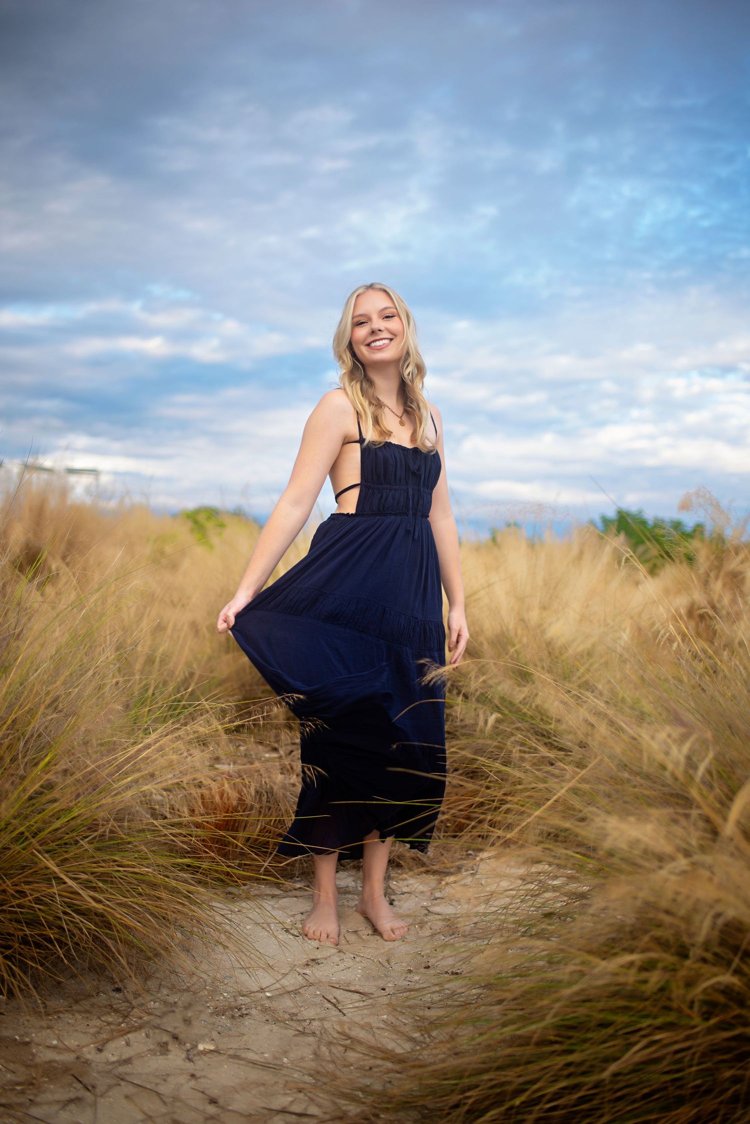 Smiling blonde woman in navy blue maxi dress standing barefoot on sandy beach path surrounded by tall golden grass.