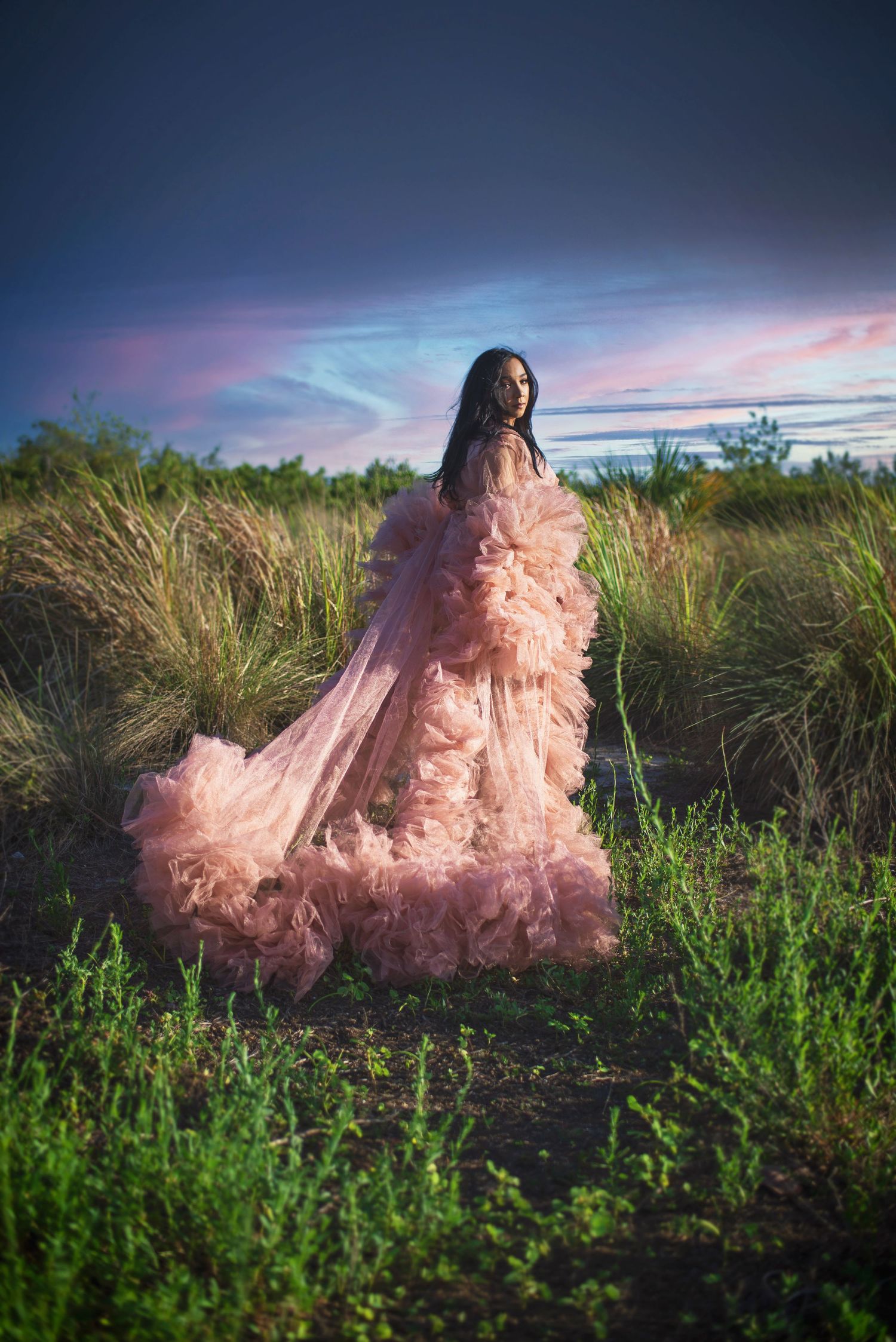 Pregnant woman in flowing pink tulle gown standing in lush green field at dramatic sunset.