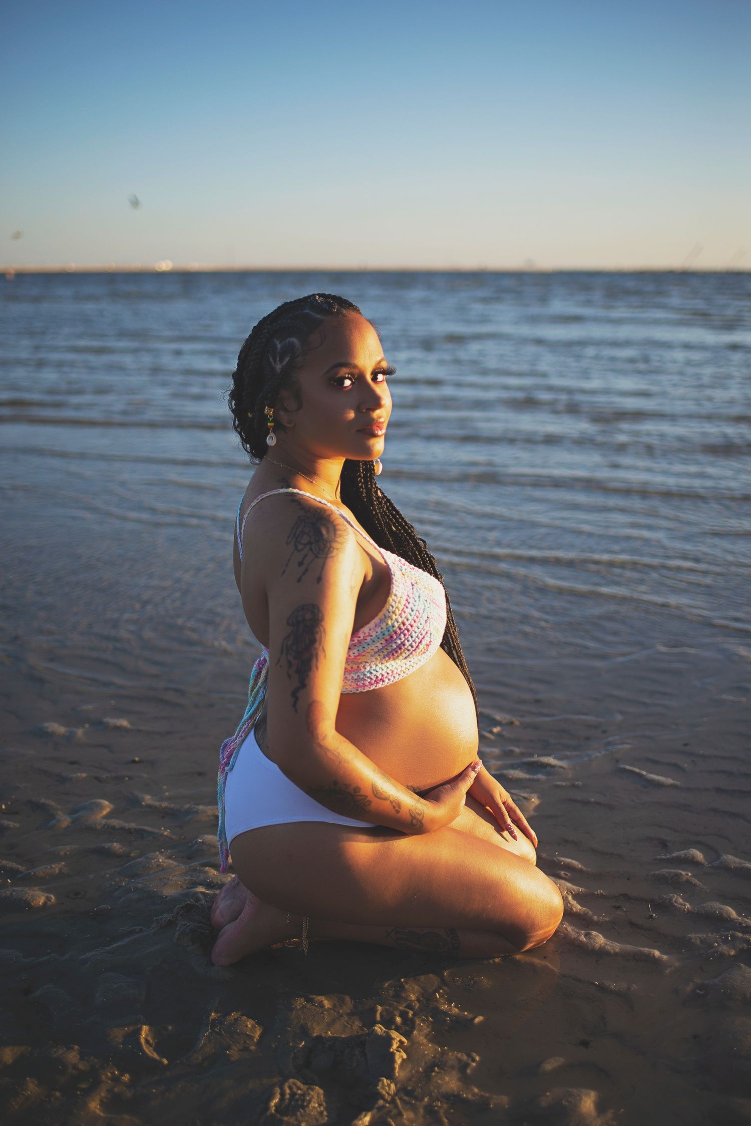 Pregnant woman in white swimsuit sitting on beach sand at sunset during maternity session.