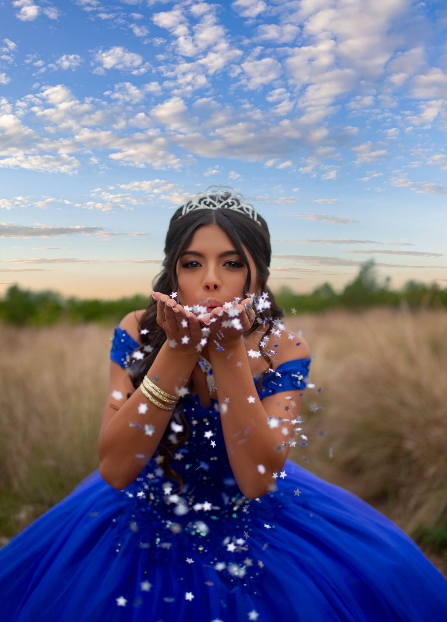Teen in royal blue ball gown and tiara blows star confetti from her hands in a scenic field.