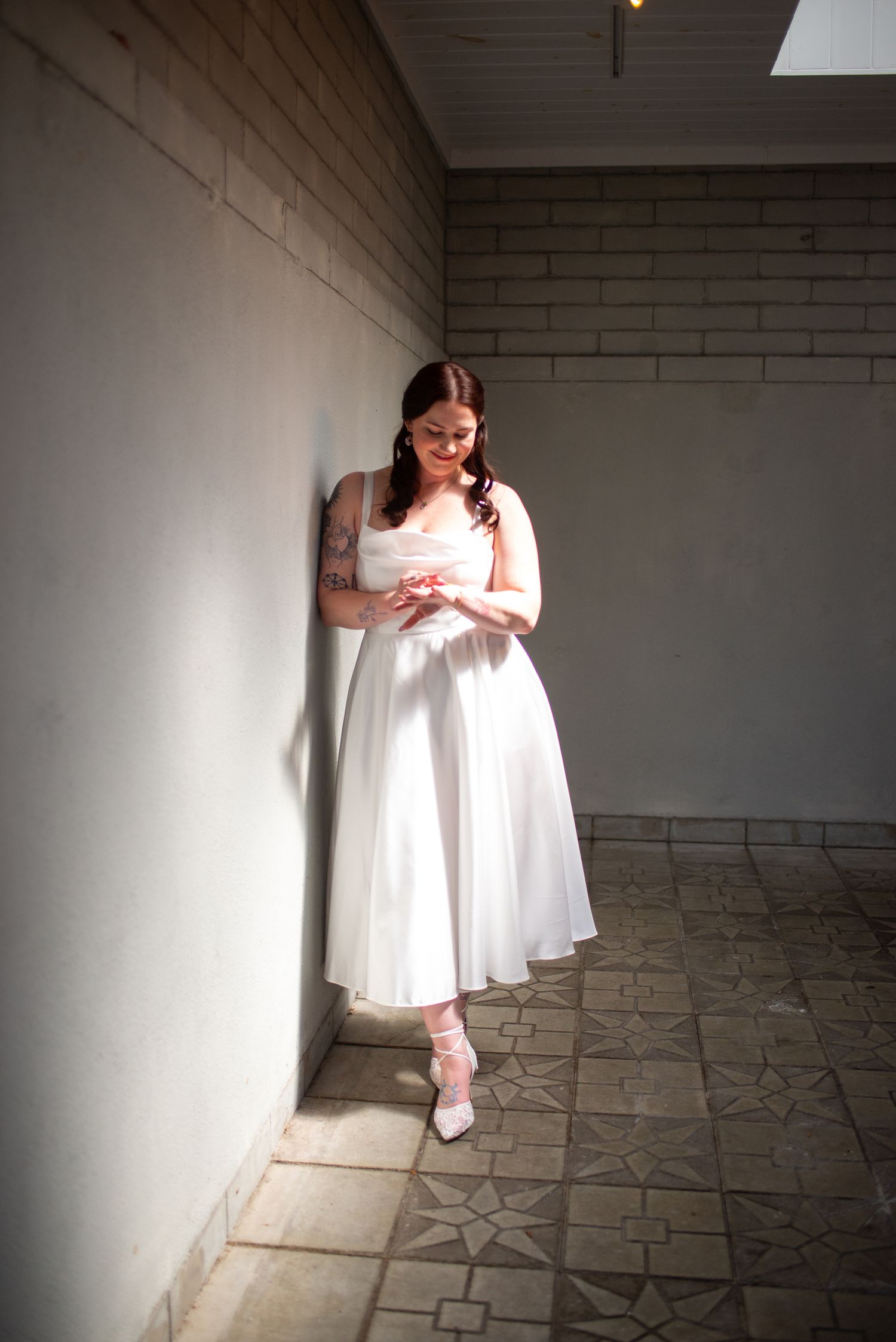 Bride in elegant white dress leans against wall in sunlit corridor with patterned tile floor.