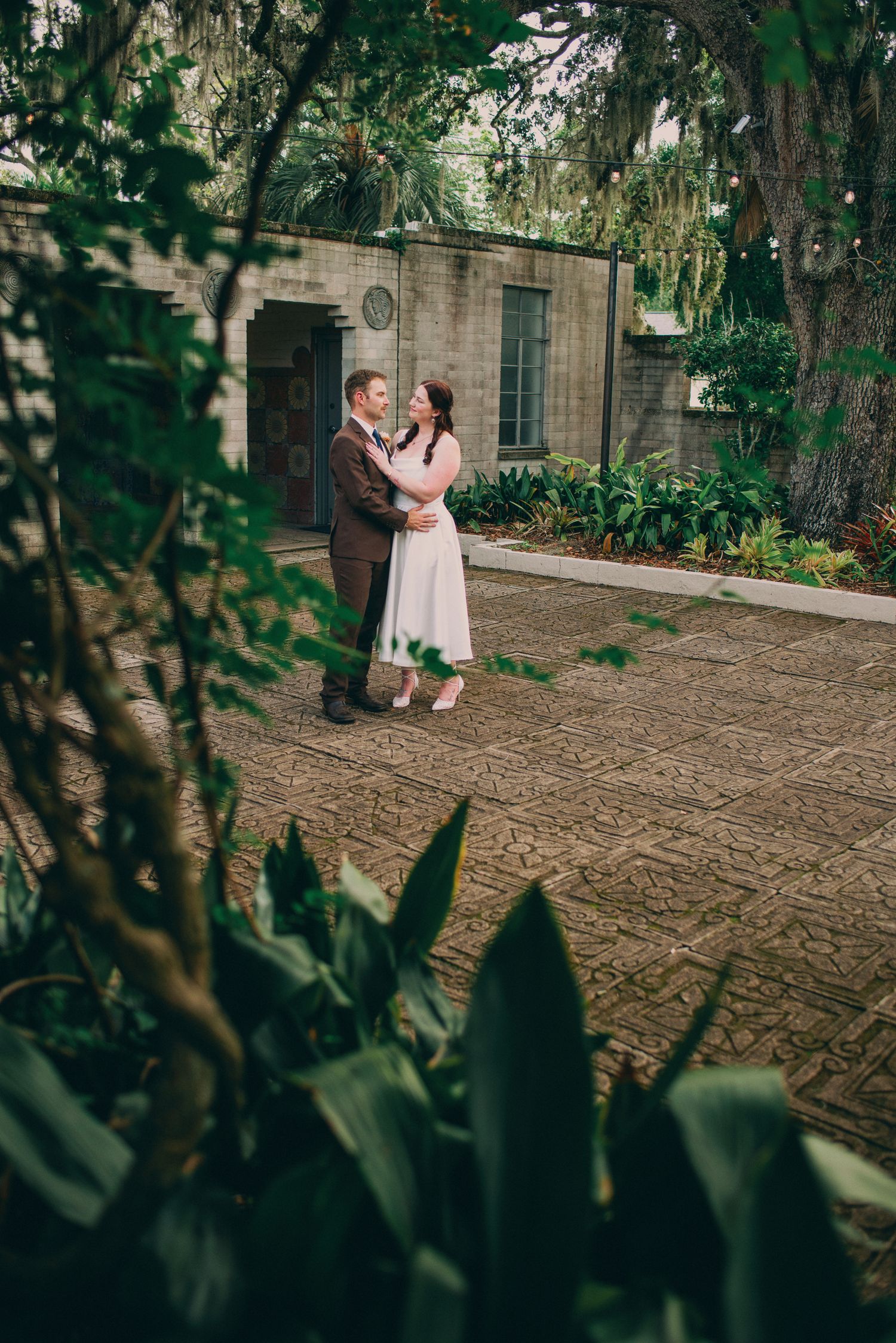 A bride and groom embrace in a lush courtyard surrounded by trees and stone walls after their wedding.