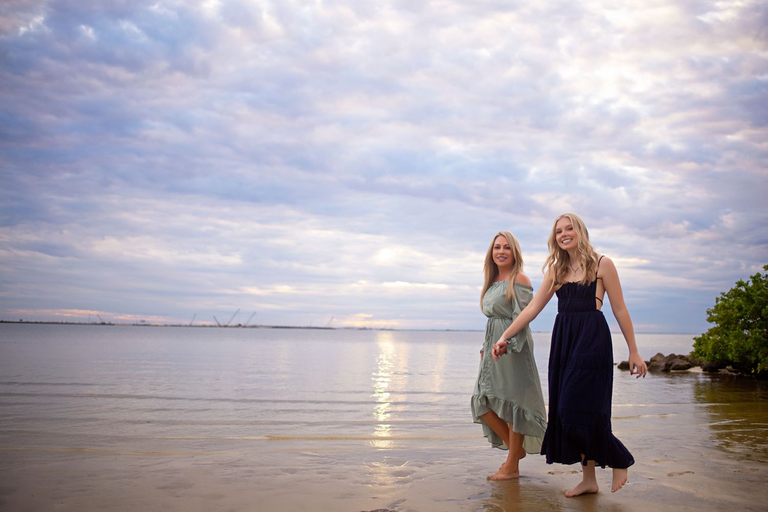 Two women in elegant dresses walking barefoot through shallow water on a serene beach at sunset.