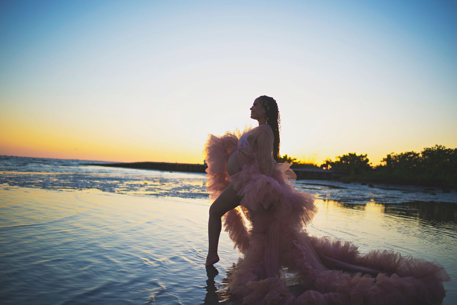 Woman in voluminous pink tulle gown posing on a calm beach shoreline during a breathtaking golden sunset.